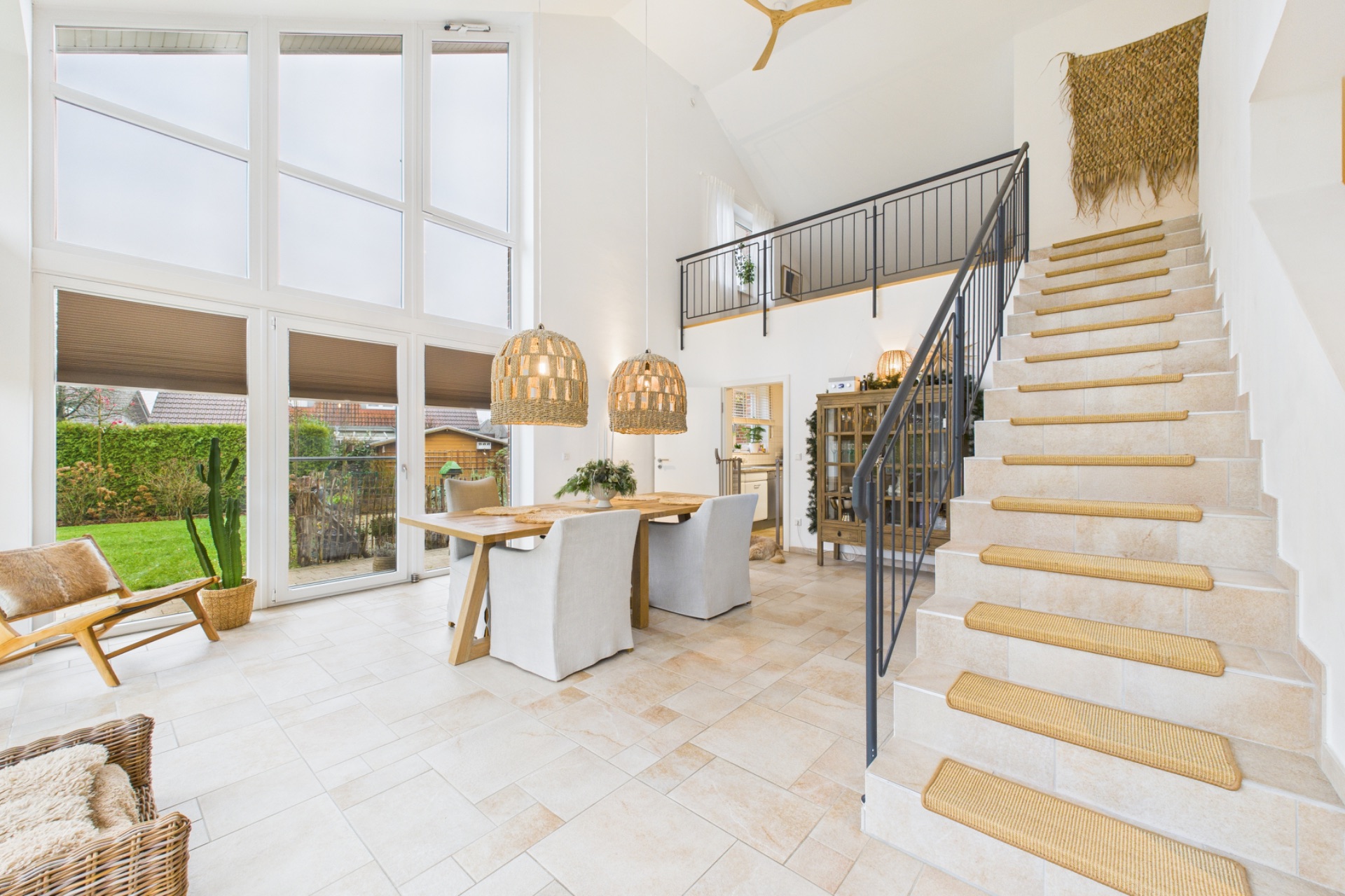 Helles Wohnzimmer mit großem Fenster, Esstisch, Holzstühlen und Treppe mit Teppichstufen.