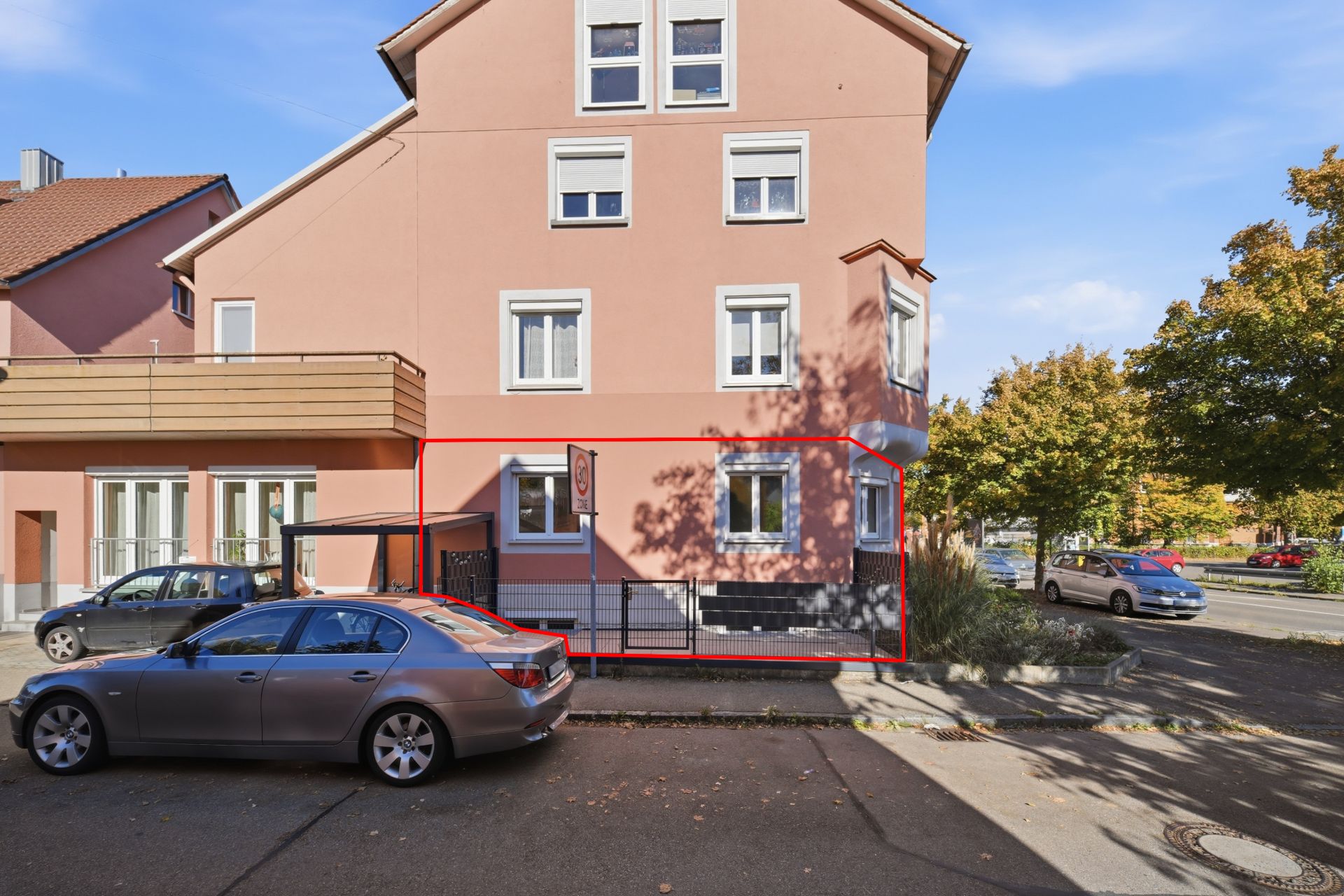 Rosa Wohnhaus an einer Straßenecke mit geparkten Autos und Bäumen im Hintergrund.