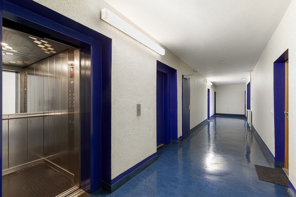 Hausflur mit blauem Boden, offenen Aufzug und mehreren Wohnungstüren.