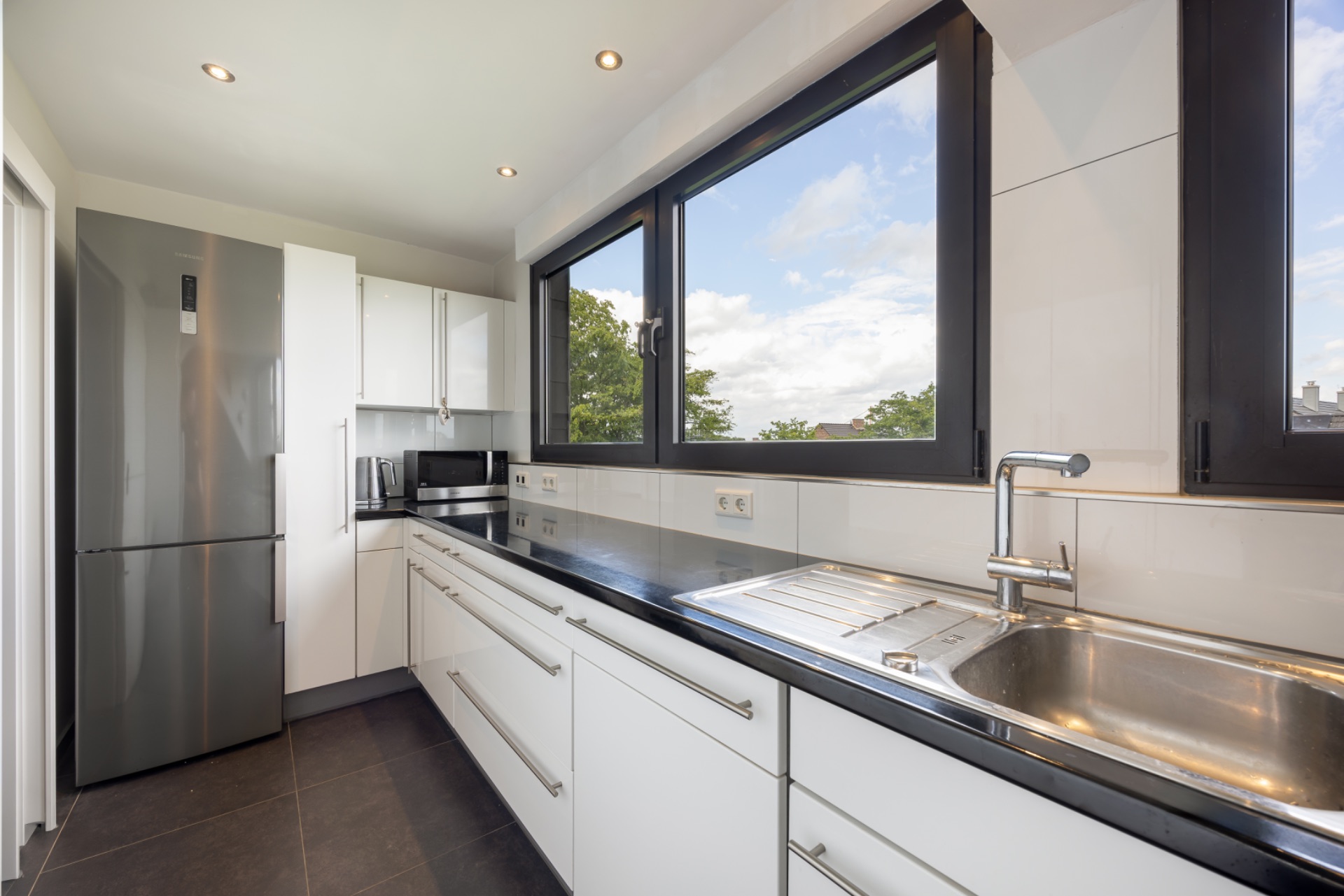 Moderne Küche mit weißen Schränken, großem Fenster, Edelstahlspüle und Kühlschrank.