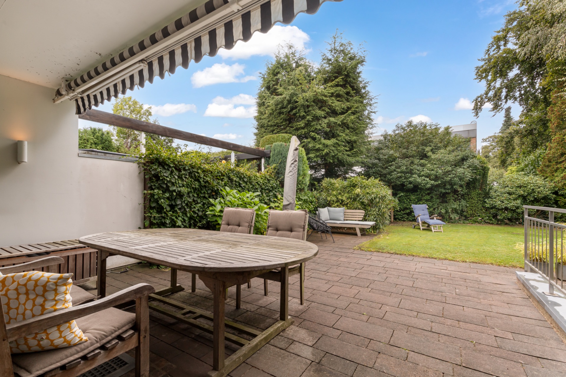 Überdachte Terrasse mit Holztisch, Stühlen und Blick auf einen Garten mit Liegestühlen.