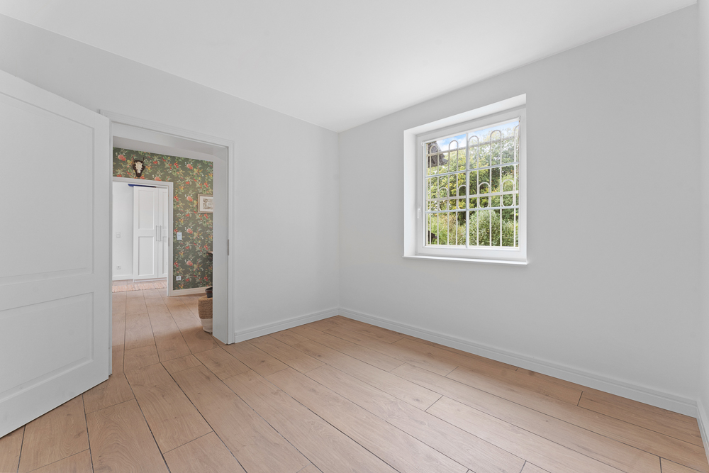 Heller leerer Raum mit Holzfußboden, weißer Wand, Fenster mit Gitter und geöffneter Tür.