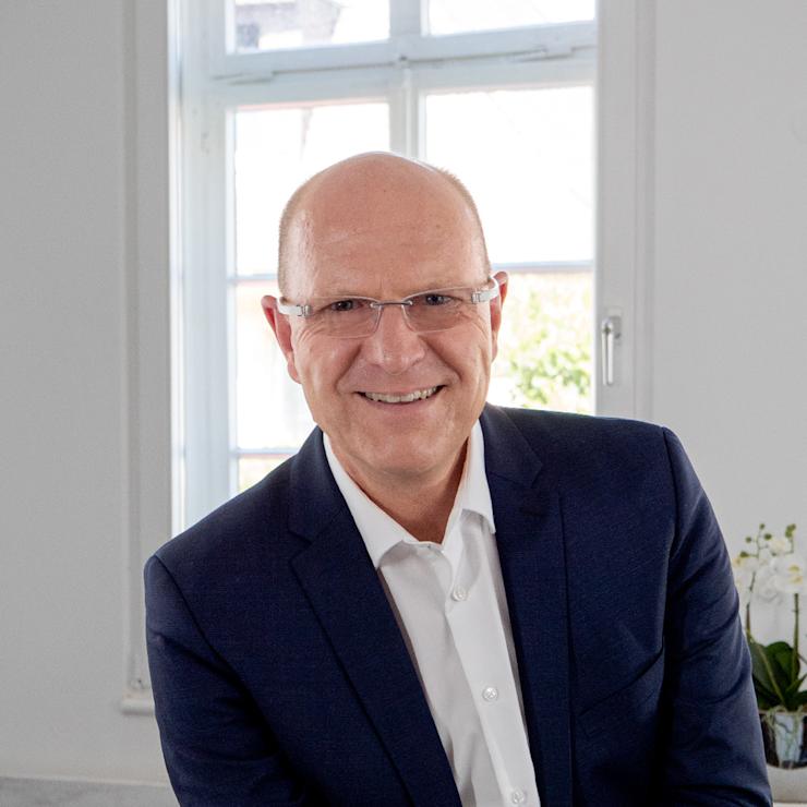 Mann mit Brille im Anzug steht vor einer Holztreppe.