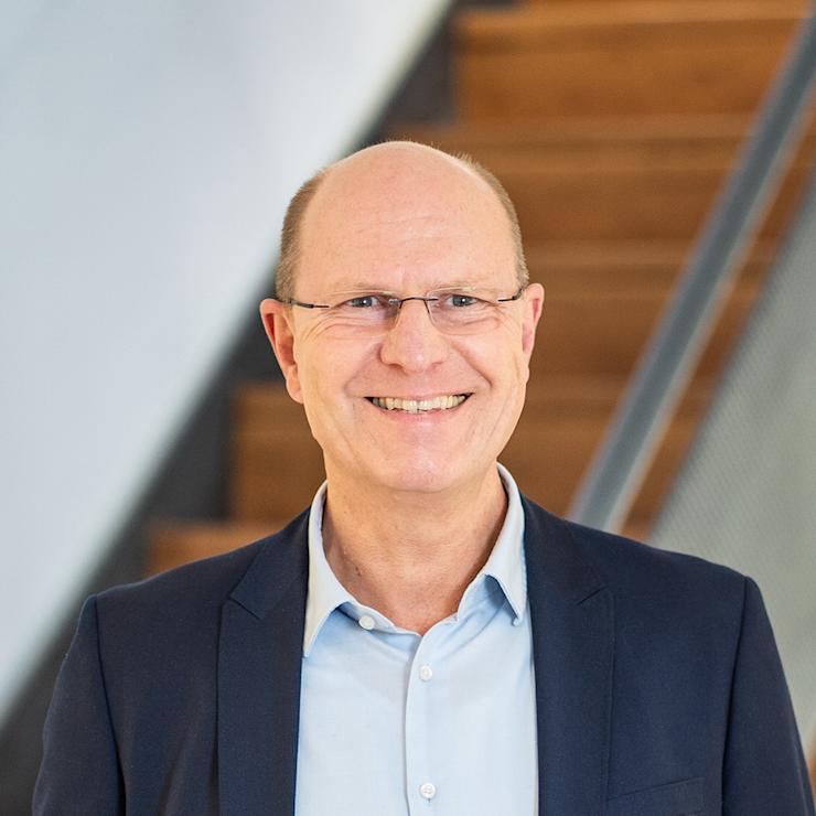 Mann mit Brille im Anzug steht vor einer Holztreppe.