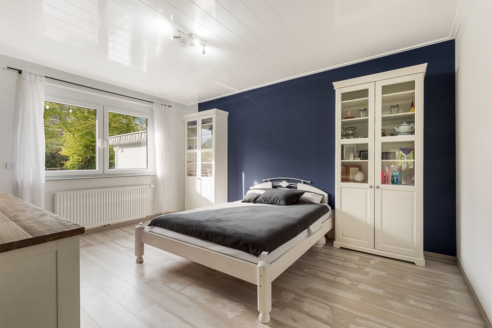 Helles Schlafzimmer mit großem Fenster, Doppelbett und zwei weißen Vitrinenschränken vor blauer Wand.