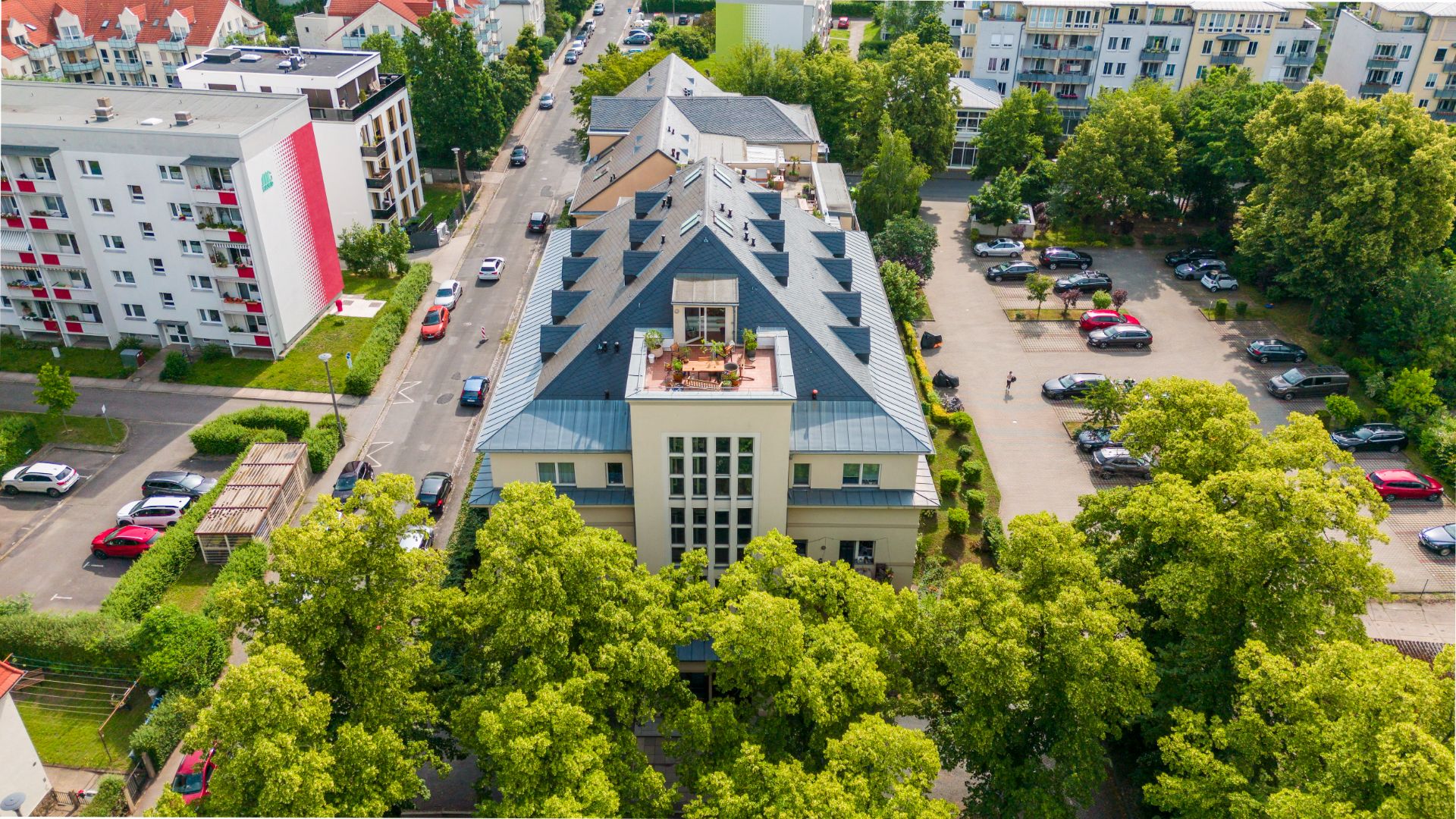 Luftaufnahme eines Wohngebäudes mit Dachterrasse, umgeben von Bäumen und Parkplätzen.
