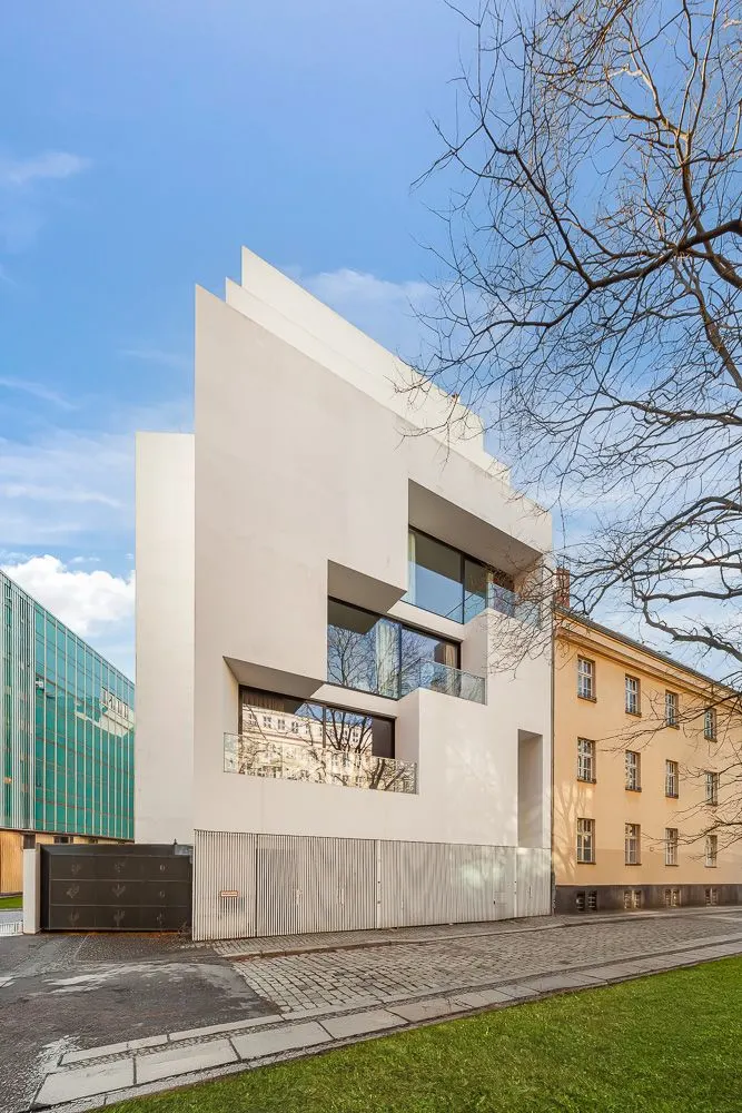 Modernes weißes Wohngebäude mit großen Fenstern und geometrischem Design, daneben ein älteres gelbes Haus.