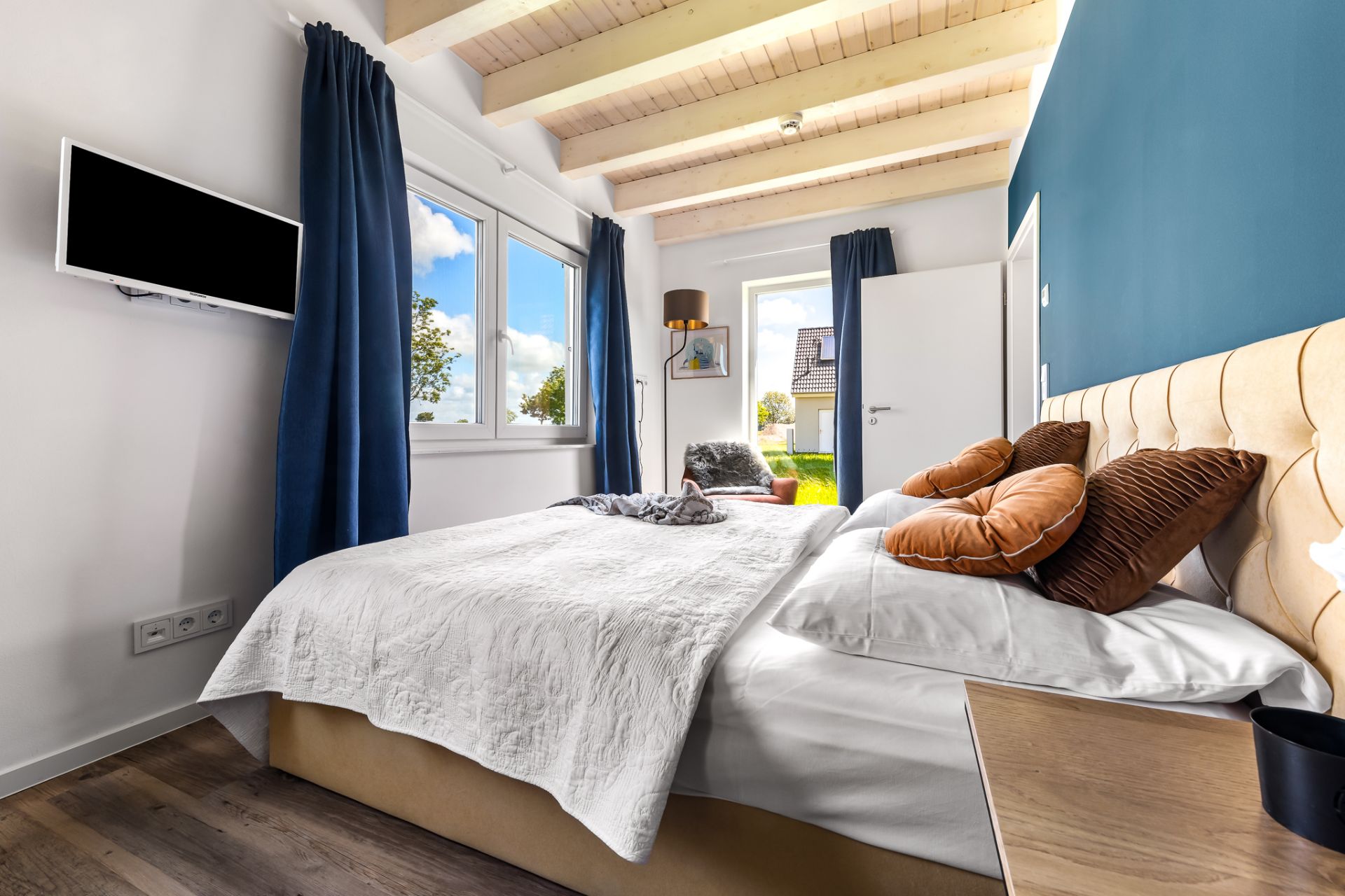 Helles Schlafzimmer mit Doppelbett, blauer Wand, TV, Fenstern und Holzbalkendecke.