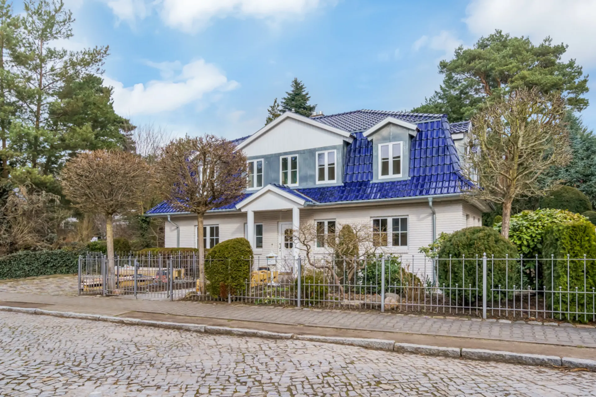 Zweistöckiges Haus mit blauem Dach, weißen Wänden, Garten und Zaun an einer gepflasterten Straße.