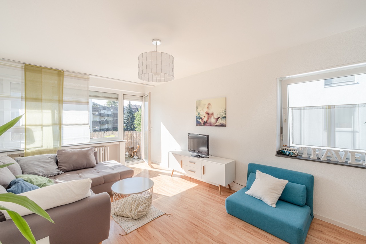 Helles Wohnzimmer mit Ecksofa, blauem Sessel, TV, Couchtisch und großen Fenstern.