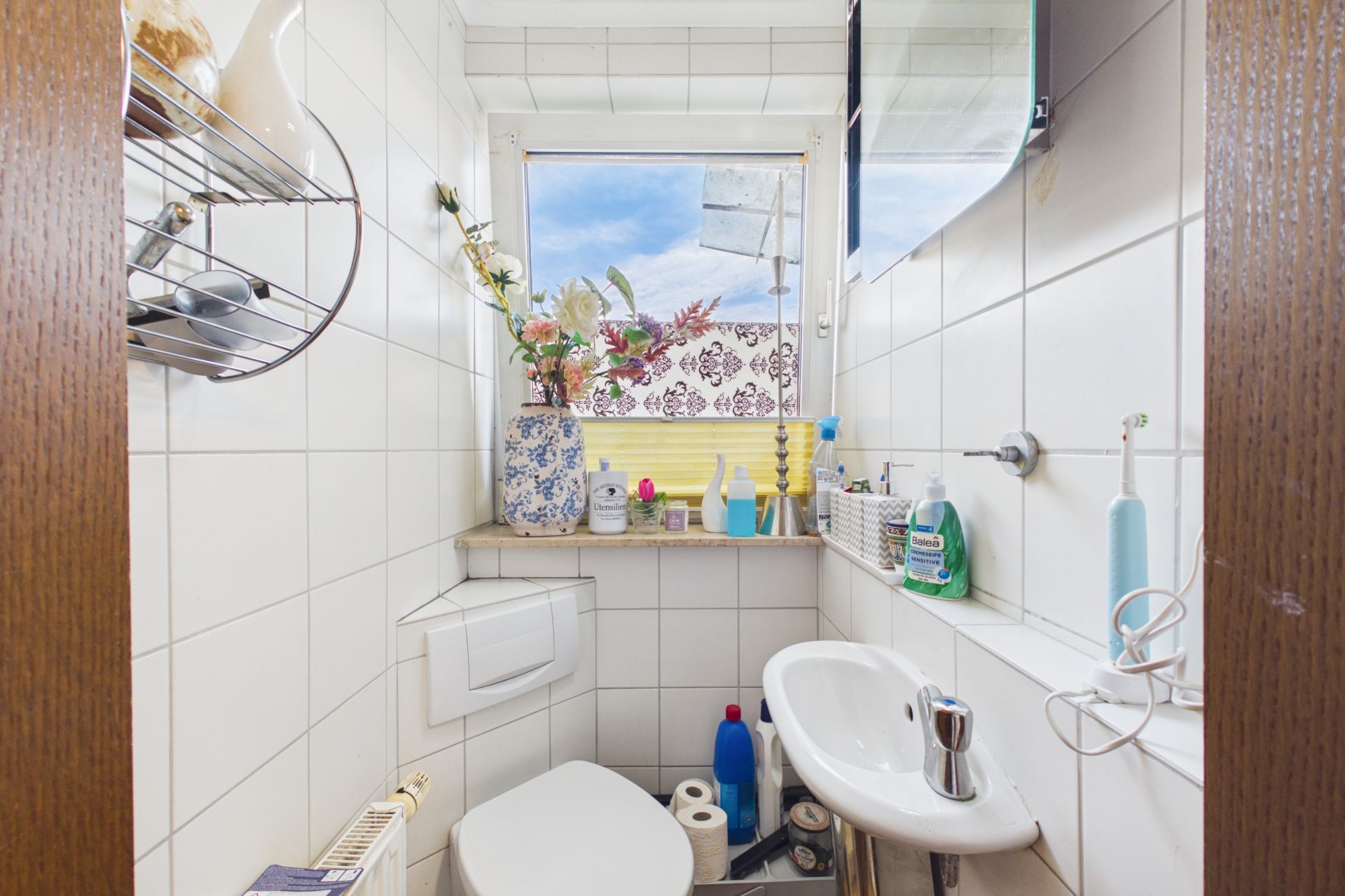 Kleines Badezimmer mit WC, Waschbecken, Zahnputzbecher und Fenster. Blumen in einer Vase auf der Fensterbank.