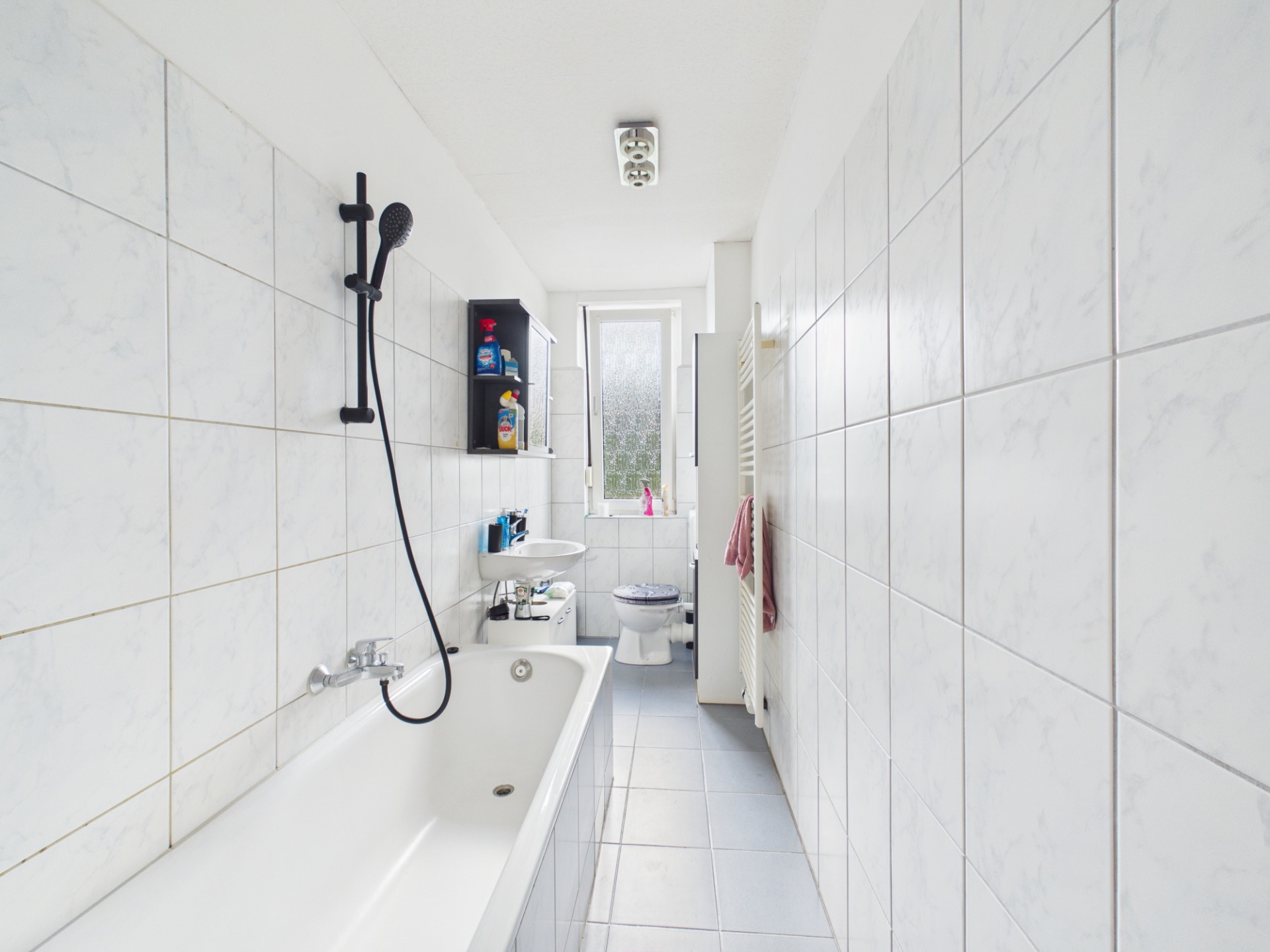 Helles Badezimmer mit Badewanne, Waschbecken, WC und Fenster, weiße Fliesen an Wänden und Boden.