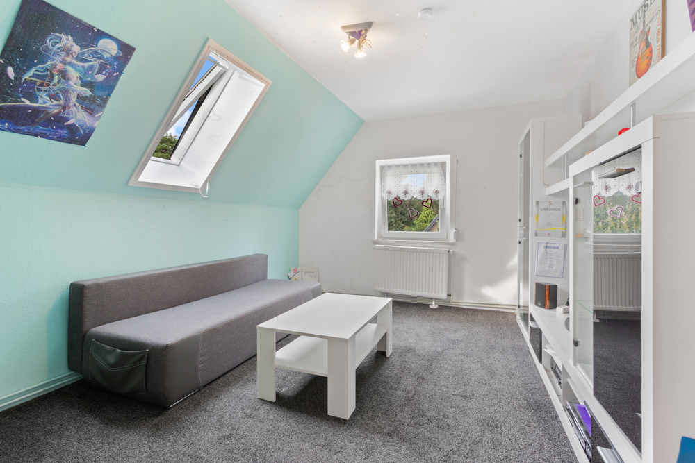 Helles Zimmer mit grauem Sofa, weißem Tisch, Regal und zwei Fenstern auf Teppichboden.