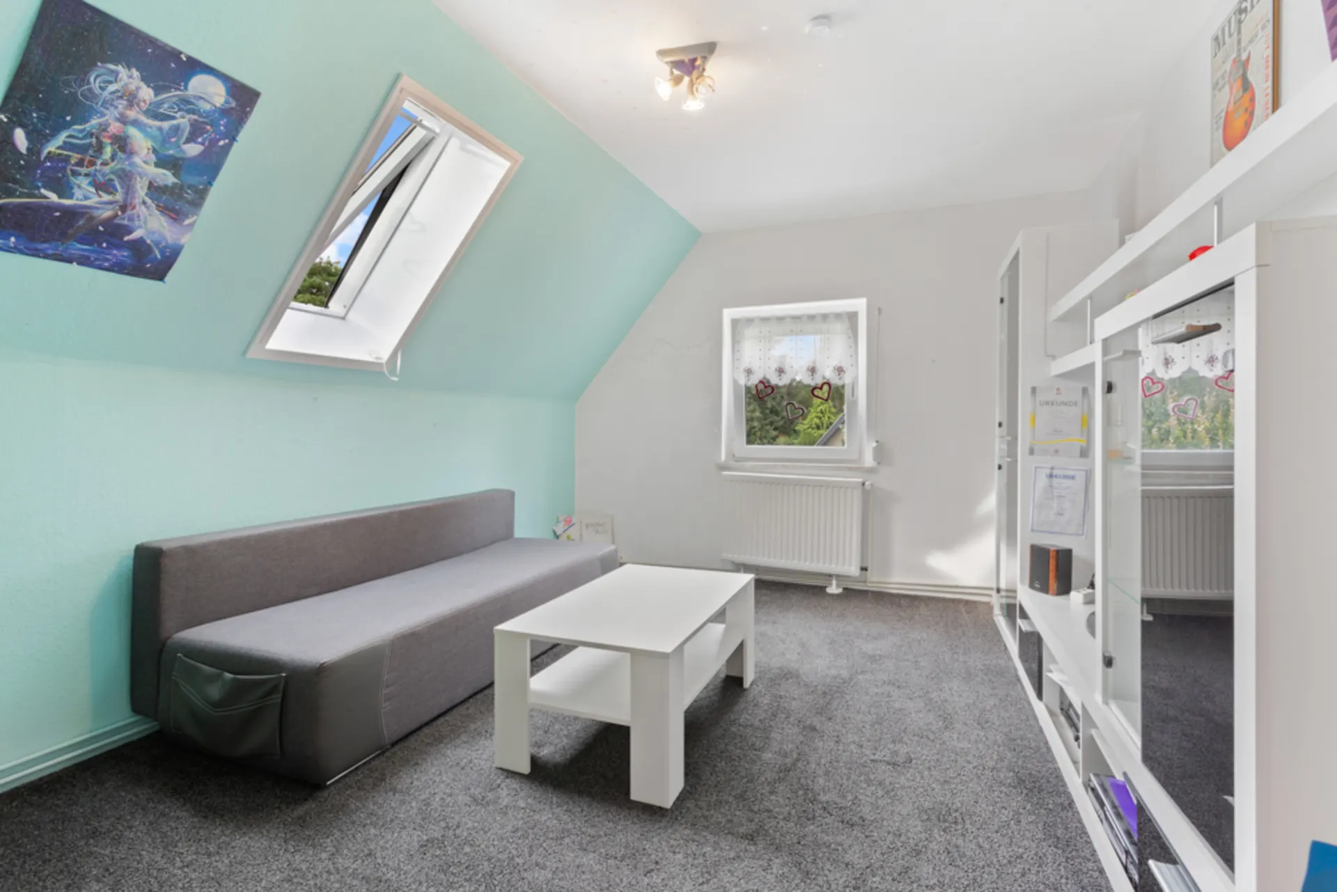 Helles Zimmer mit grauem Sofa, weißem Tisch, Regal und zwei Fenstern auf Teppichboden.