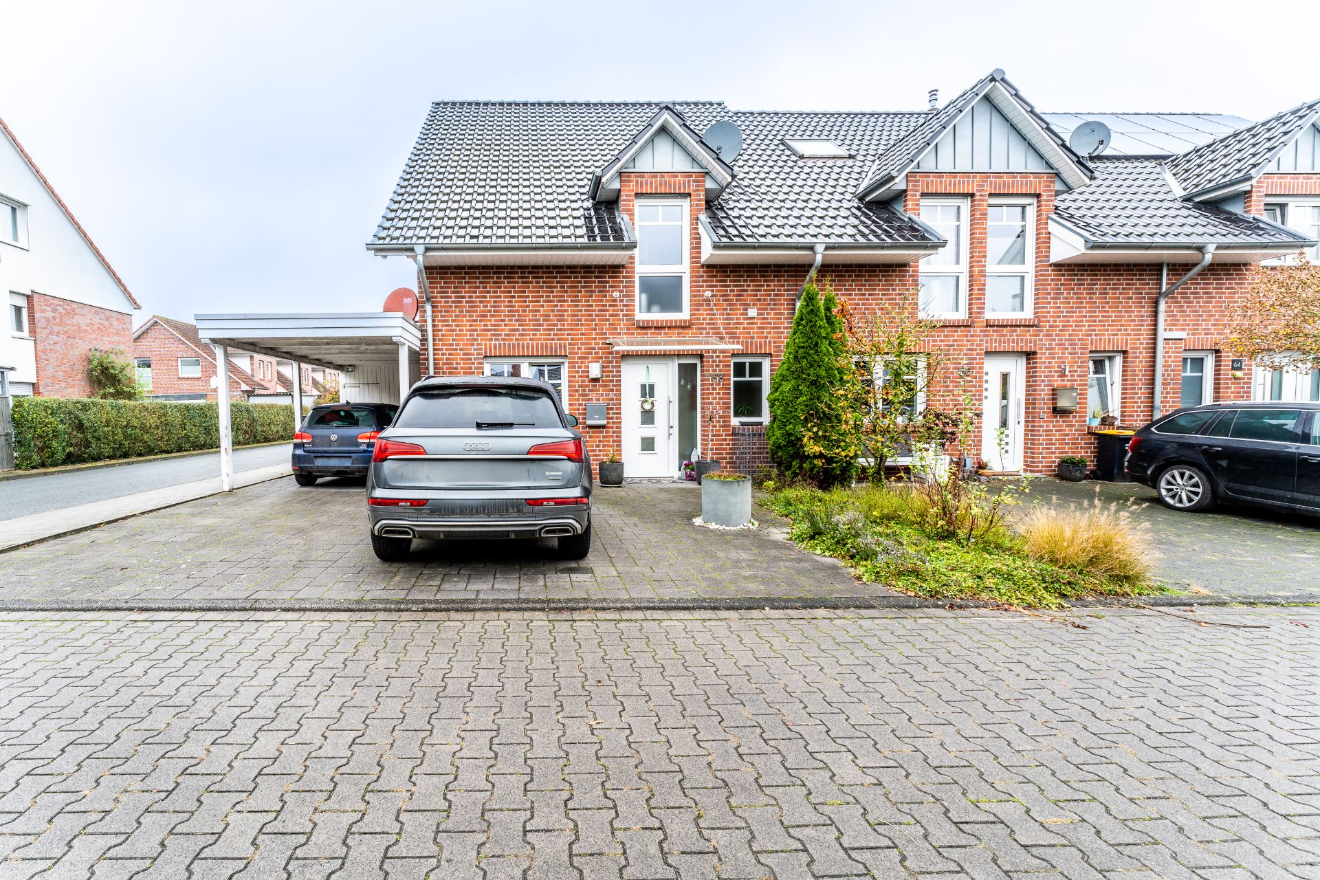 Zweistöckiges Reihenhaus aus rotem Backstein mit drei parkenden Autos und kleinem Vorgarten.