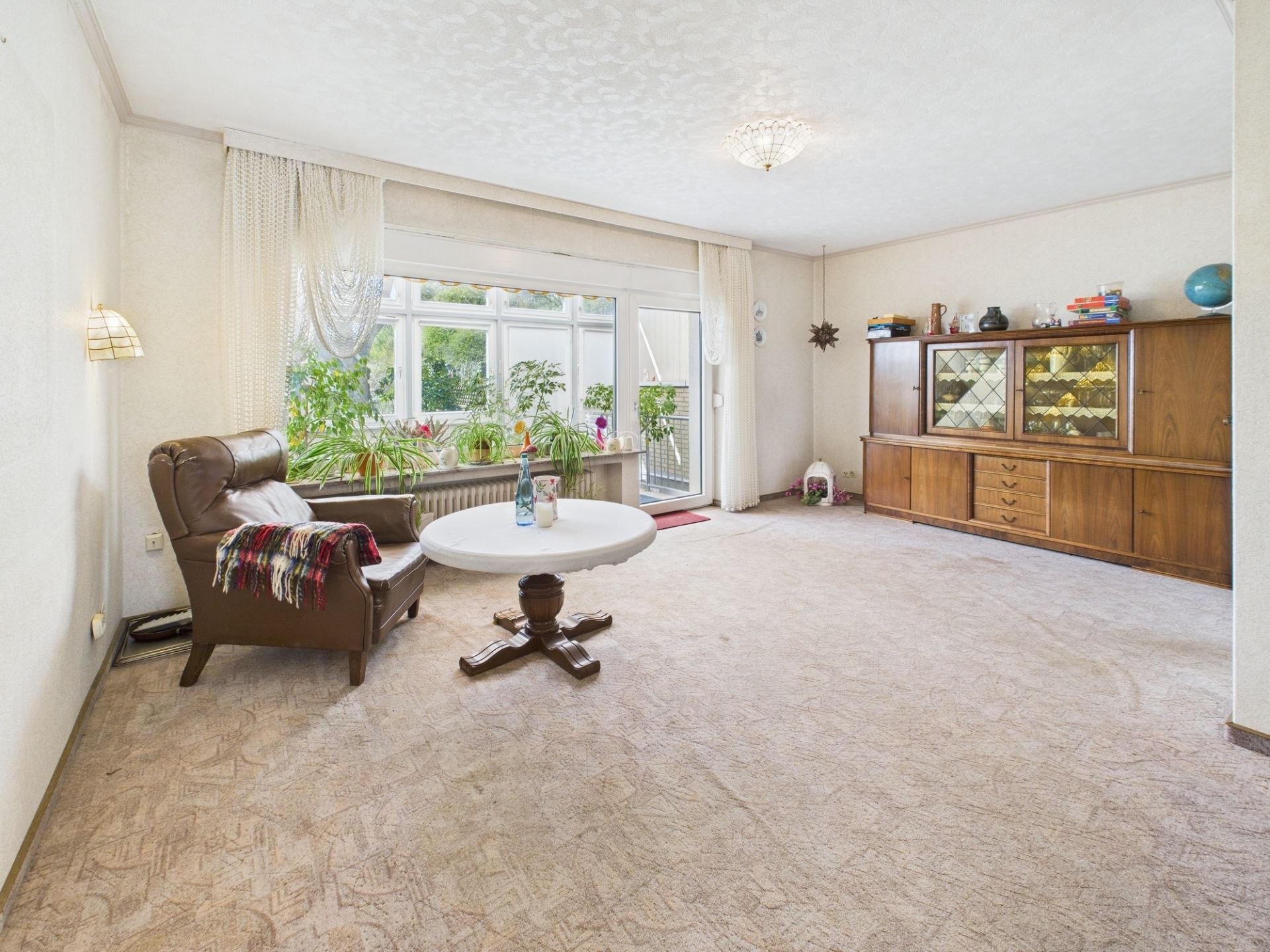 Helles Wohnzimmer mit Teppich, Sessel, rundem Tisch, Sideboard und großen Fenstern mit Blick ins Grüne.