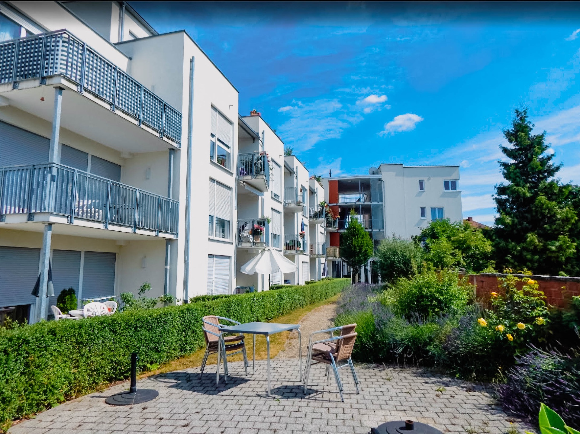 Moderne Wohngebäude mit Balkonen, Gartenweg und Sitzgruppe im Grünen bei blauem Himmel.