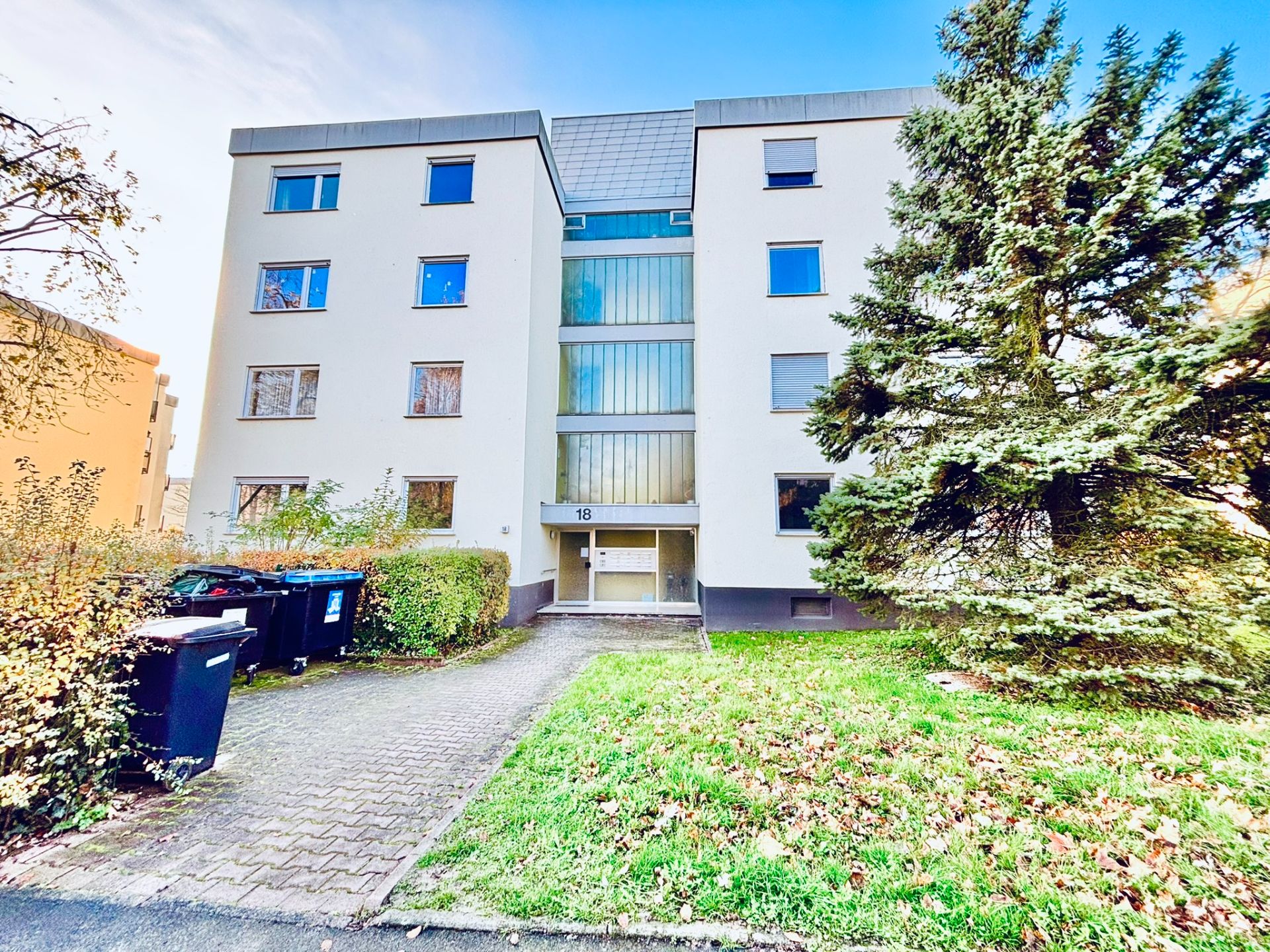 Mehrfamilienhaus mit vier Stockwerken, Eingang in der Mitte, vorne Rasen und Mülltonnen.