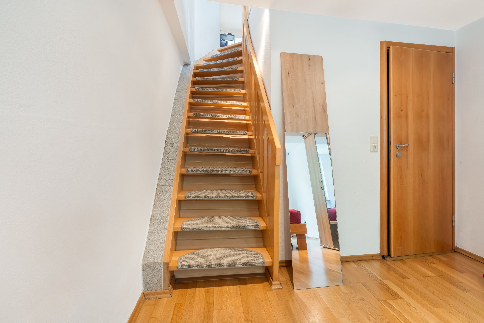 Holztreppe mit Teppichstufen, Wandspiegel und Holztür auf Parkettboden.