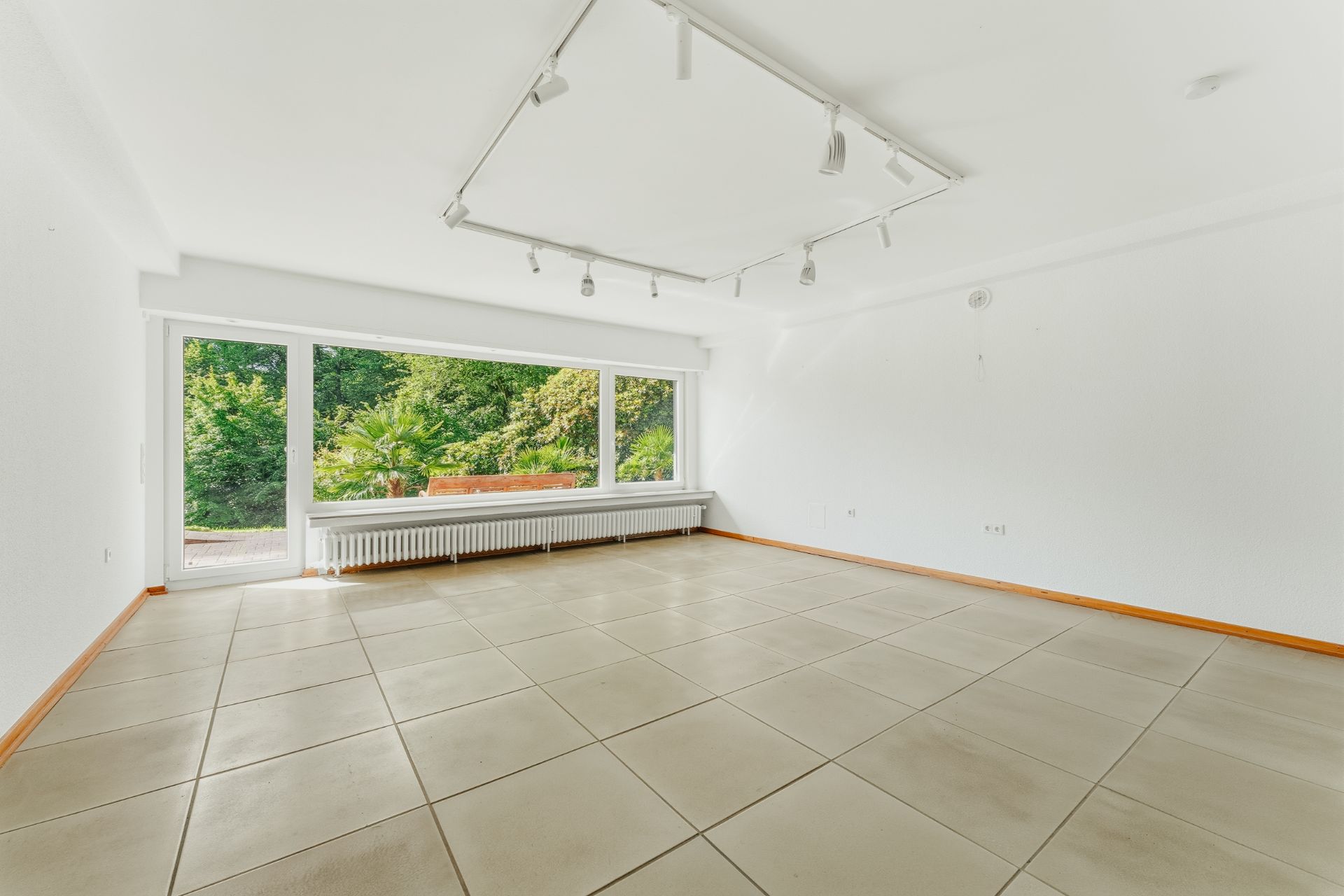 Helles, leeres Wohnzimmer mit großen Fenstern, Fliesenboden und Blick auf Garten.