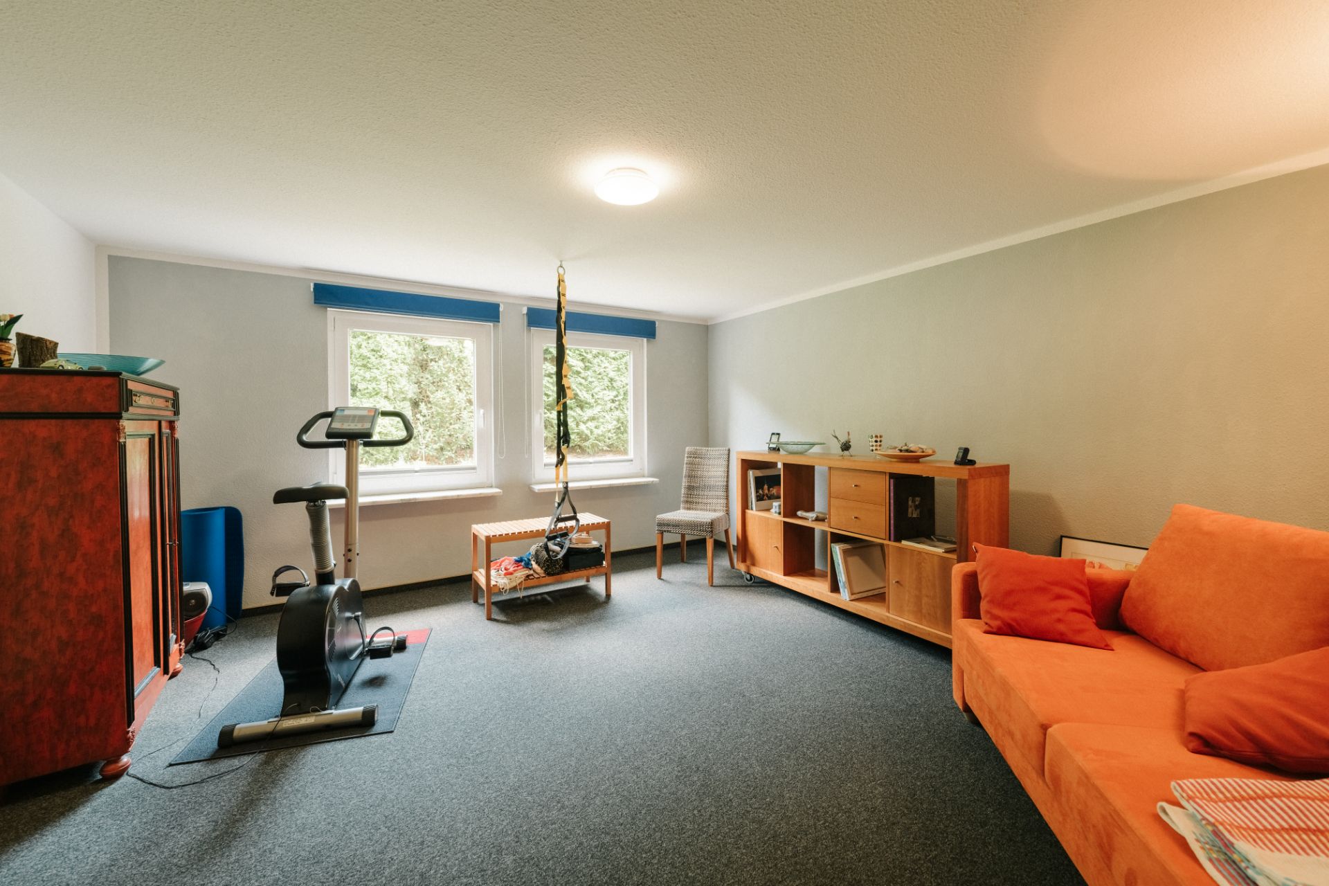 Helles Wohnzimmer mit orangem Sofa, Heimtrainer, Holzregal und zwei Fenstern.
