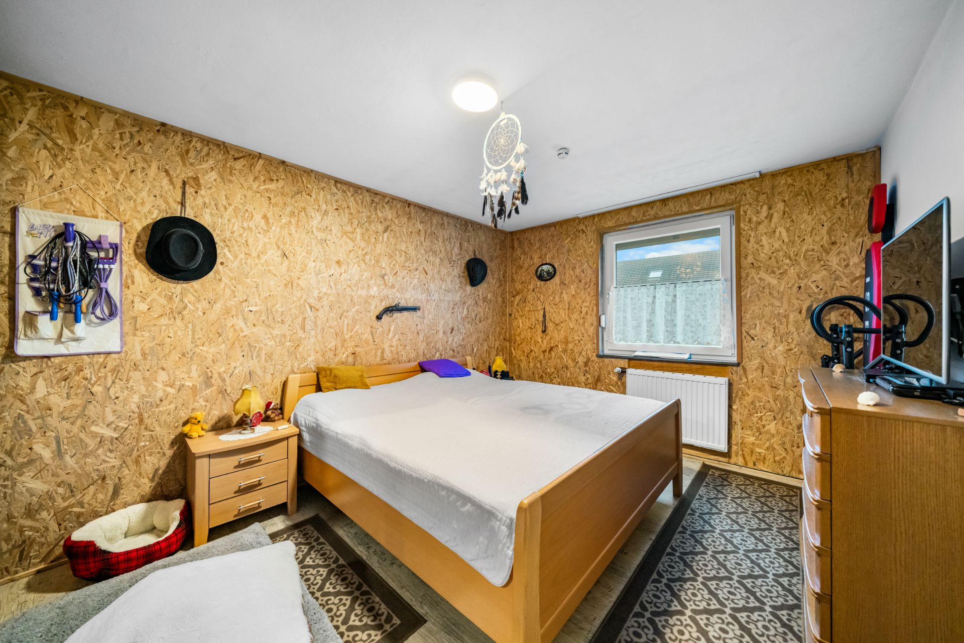 Schlafzimmer mit Holzdekor, Doppelbett, Nachttisch, Kommode und Fenster.