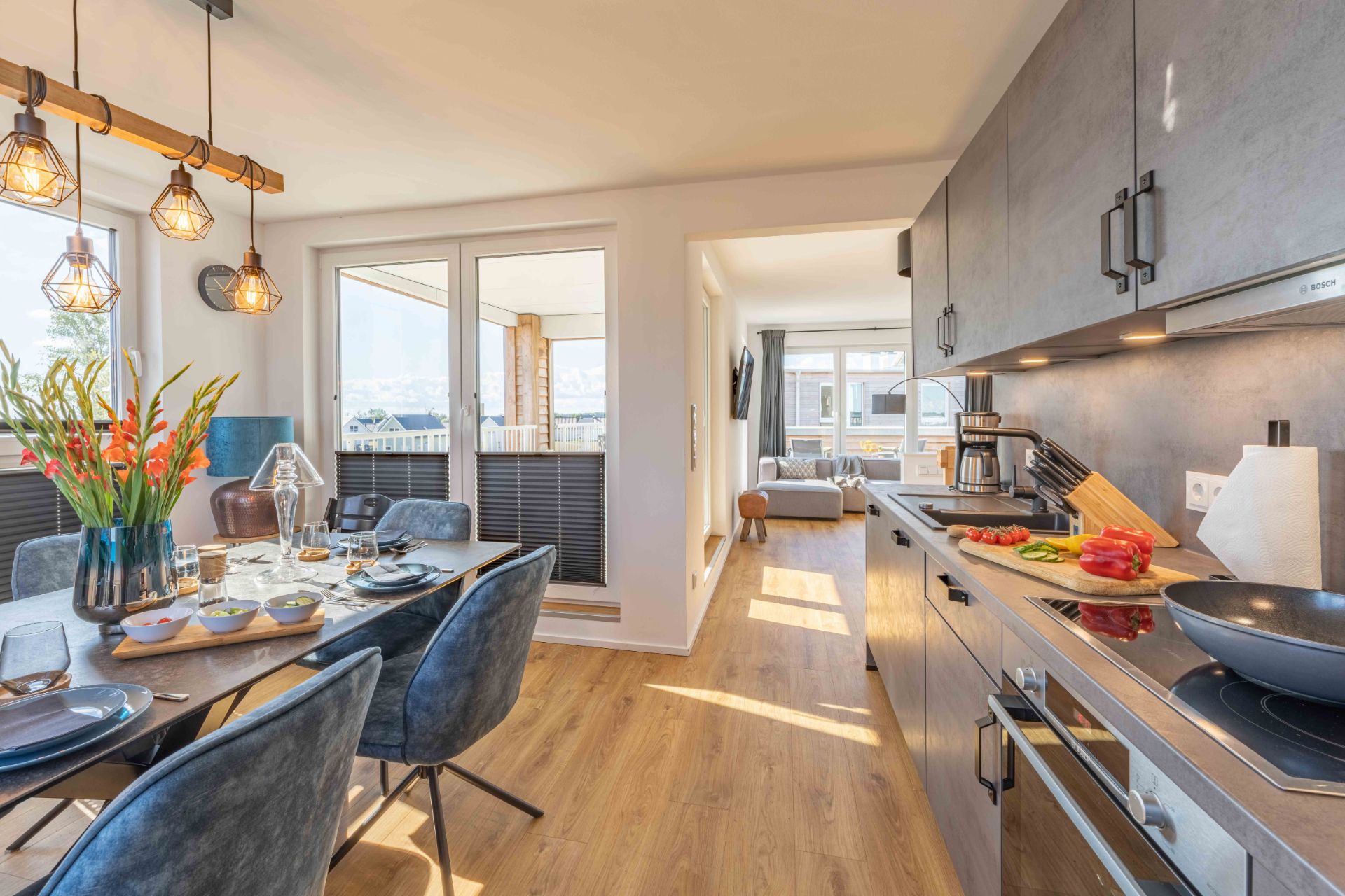 Moderne Küche mit Esstisch und Blick ins helle Wohnzimmer, große Fenster und Holzfußboden sichtbar.
