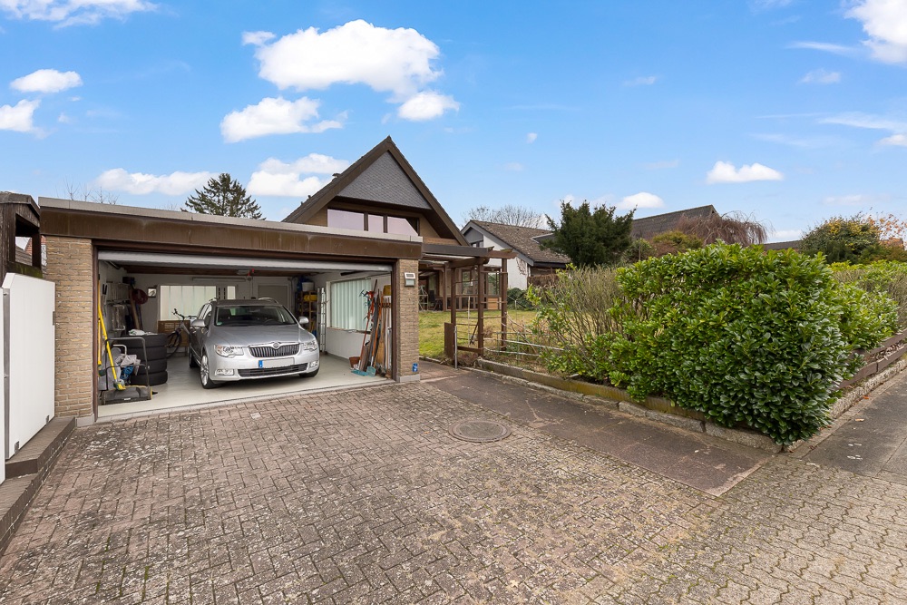 Offene Garage mit Auto und Werkzeugen, neben einem Haus mit Garten und Sträuchern.