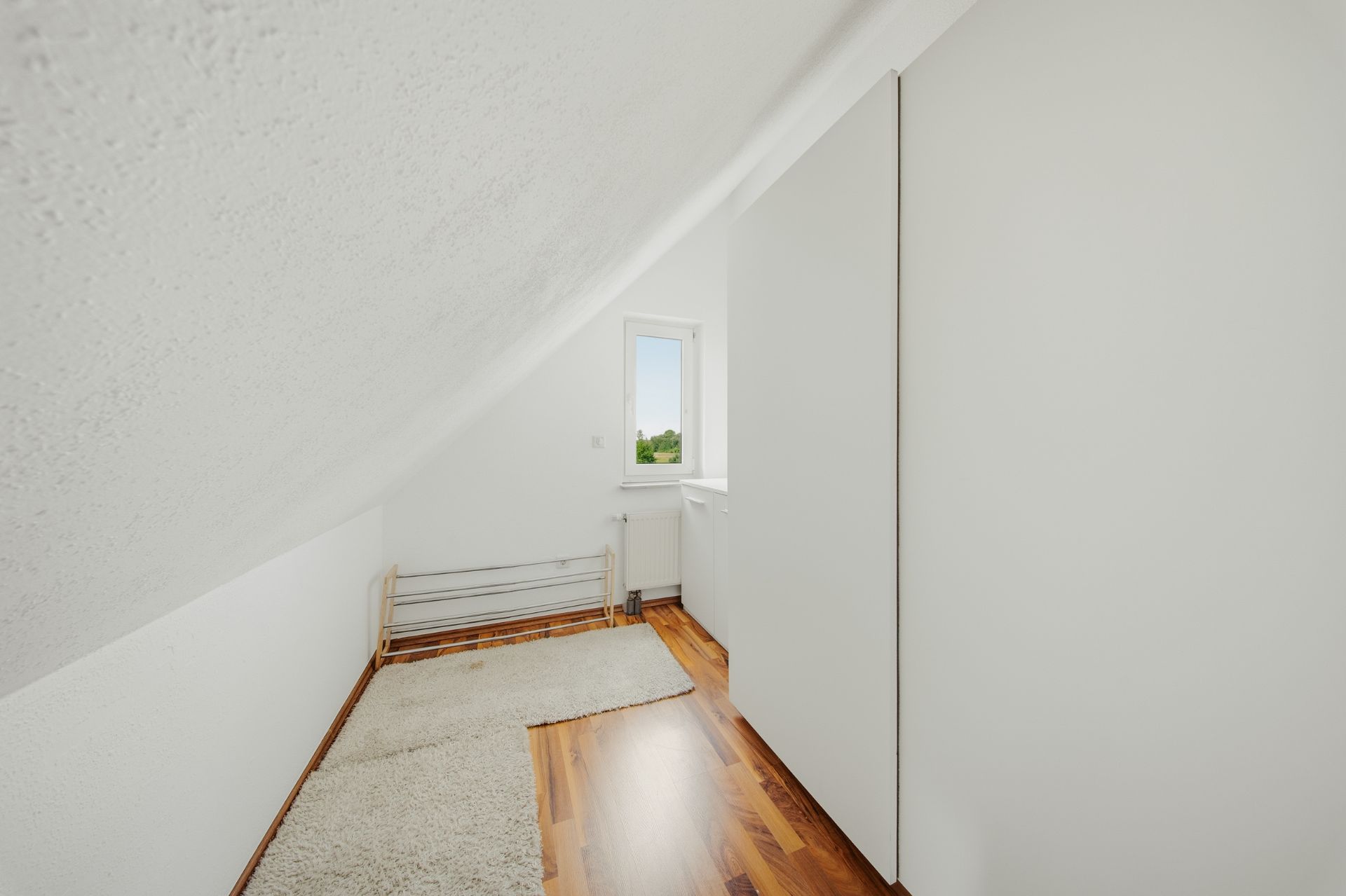 Kleiner Raum mit Dachschräge, Holzboden, Teppich, Fenster und weißem Wandschrank.