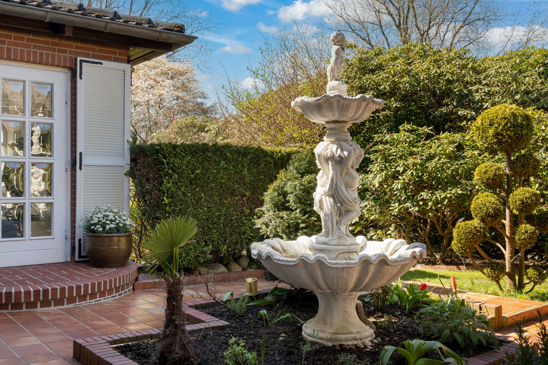 Steinerner Gartenbrunnen mit Figur neben Terrasse und Glastür, umgeben von Pflanzen und Sträuchern.