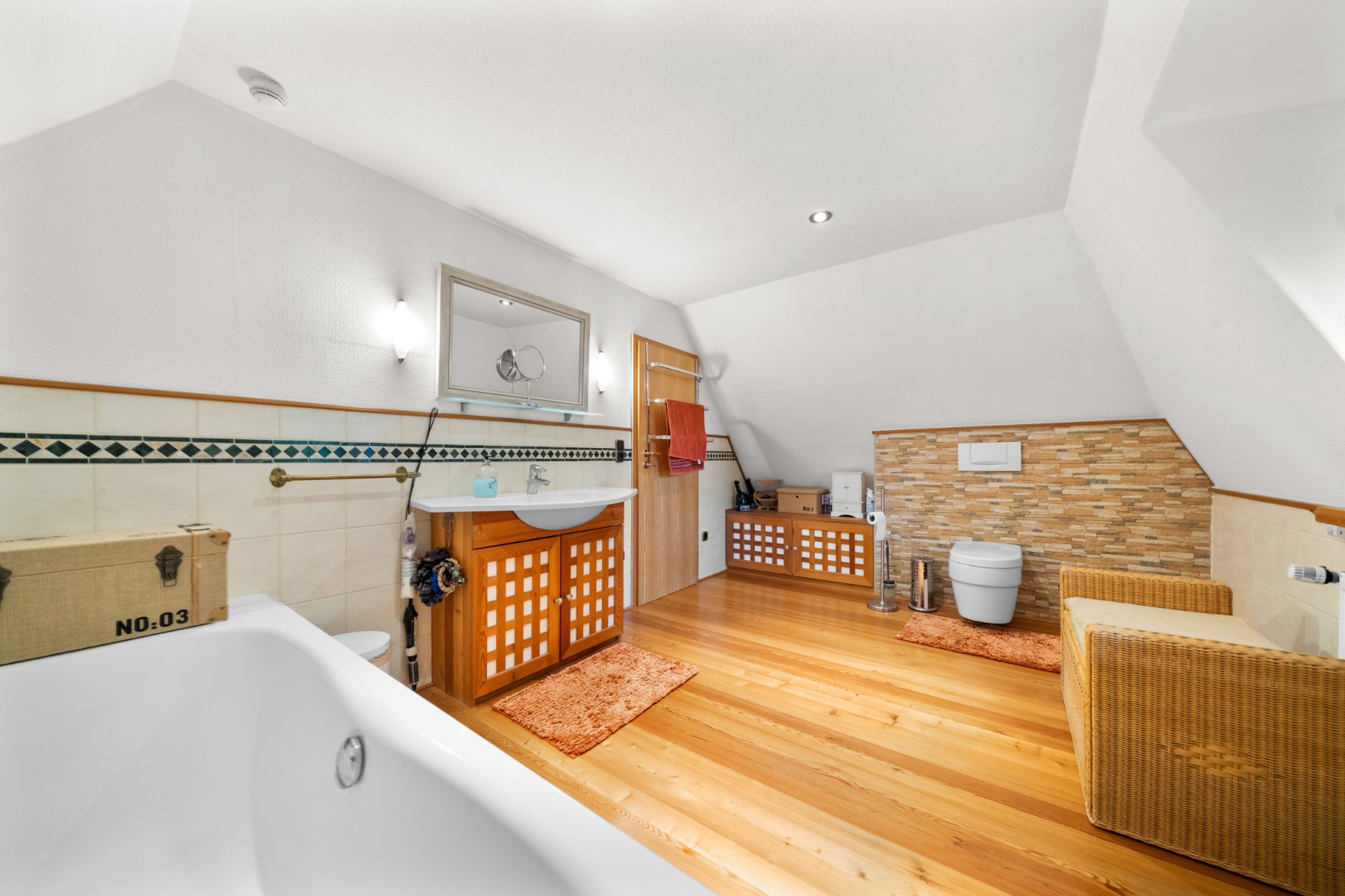 Helles Bad mit Badewanne, Waschbecken, WC, Holzfußboden und geflochtenem Sessel.