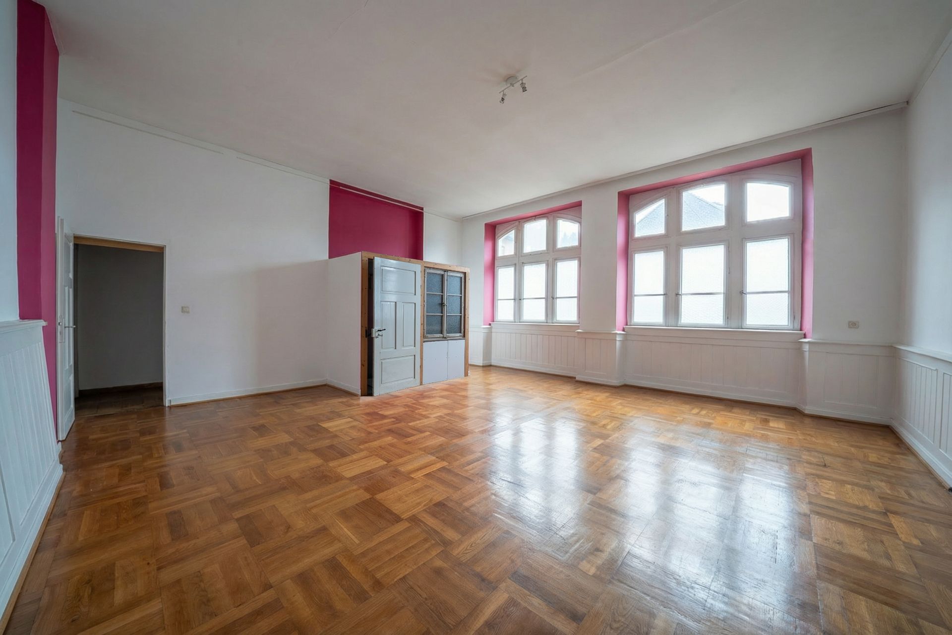 Helles, leeres Zimmer mit großen Fenstern, Parkettboden und weißen Wänden mit pinken Akzenten.