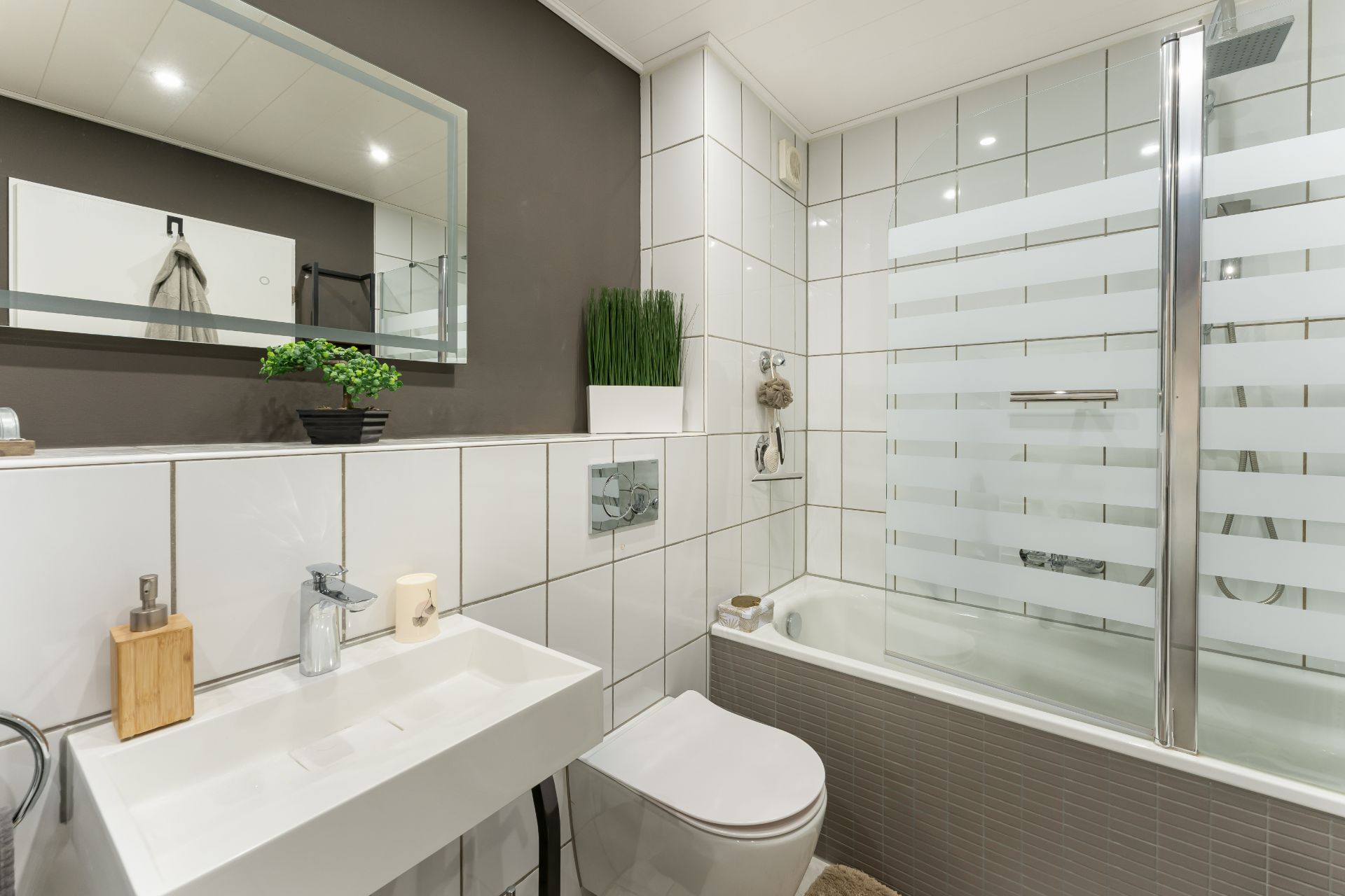 Modernes Badezimmer mit weißer Fliesenwand, Waschbecken, Toilette und Badewanne mit Glasabtrennung.
