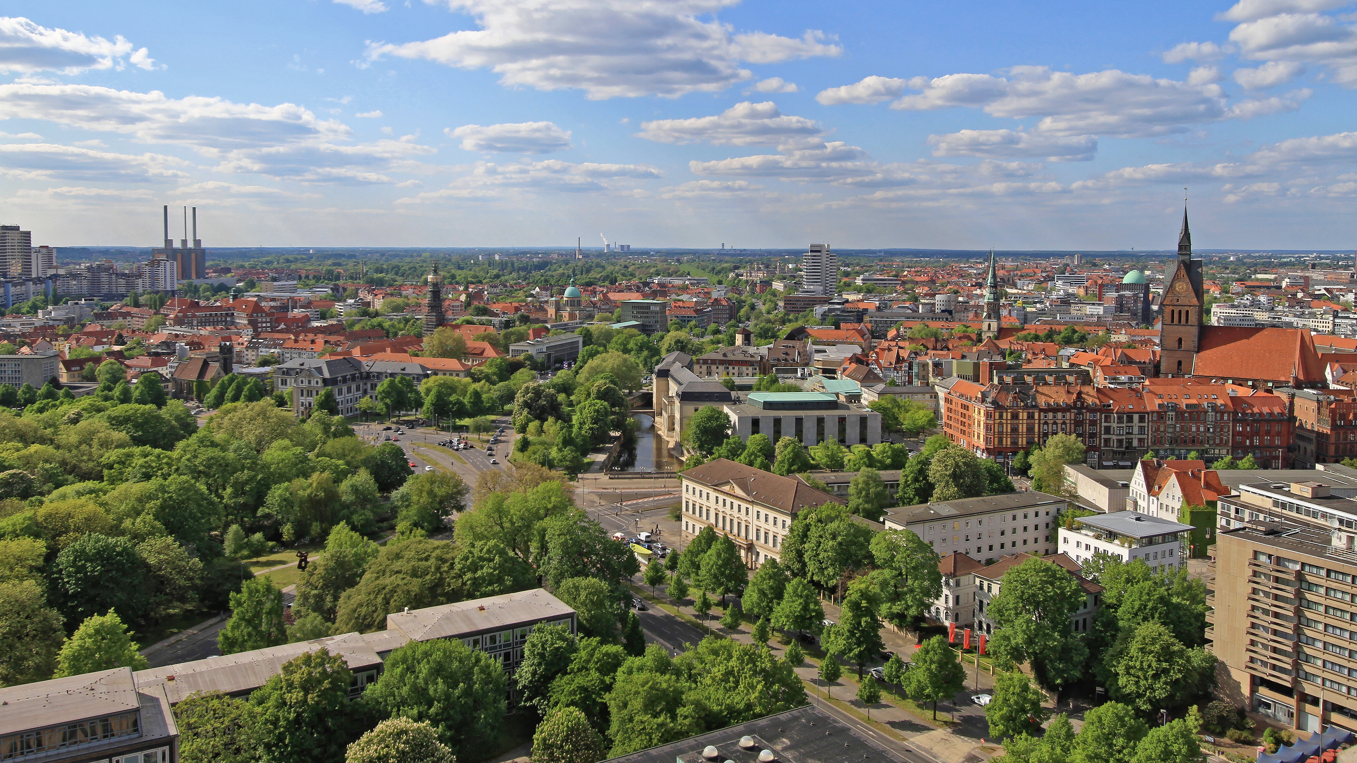 Blick über eine Stadt mit vielen Bäumen, Gebäuden und einer Kirche unter blauem Himmel.