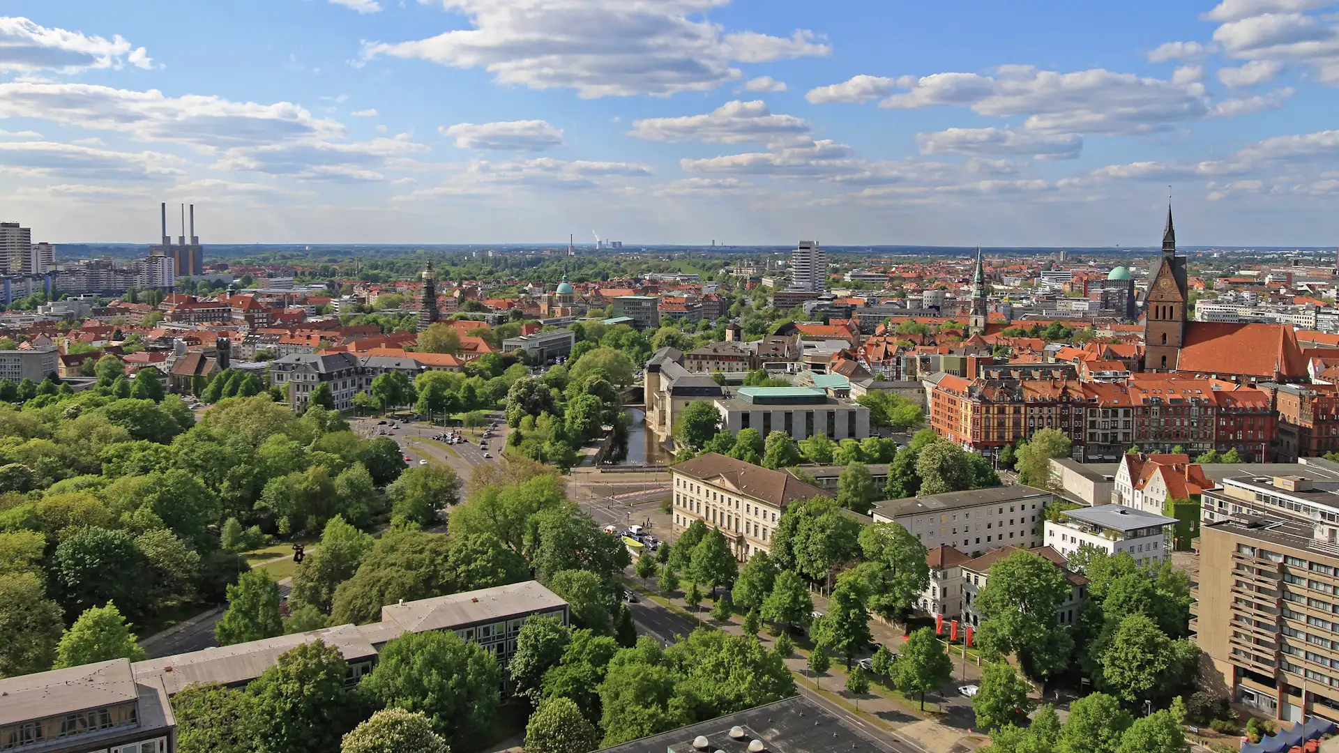 Blick über eine Stadt mit vielen Bäumen, Gebäuden und einer Kirche unter blauem Himmel.