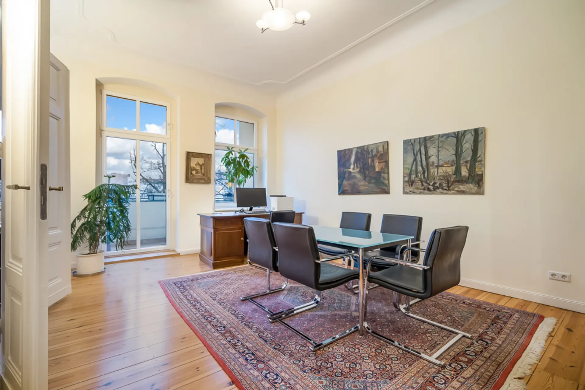 Helles Arbeitszimmer mit Glastisch, sechs Stühlen, Holzboden, Teppich, Pflanzen und Bildern an der Wand.