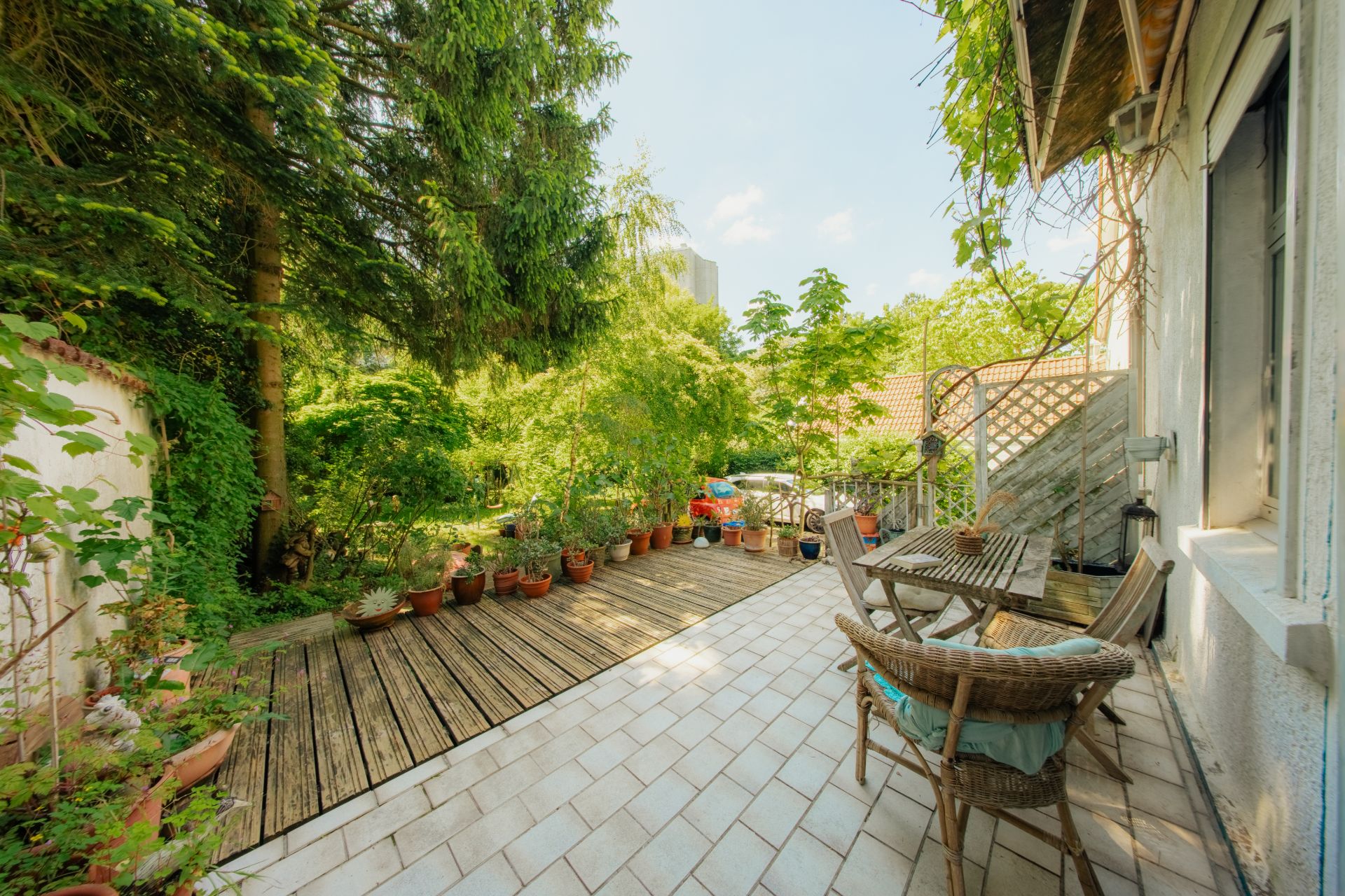 Terrasse mit Holztisch, Stühlen und vielen Blumentöpfen, umgeben von Bäumen und Pflanzen.
