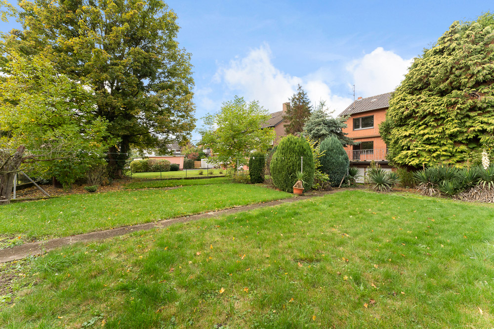 Grüner Garten mit Rasen, Bäumen und Sträuchern, im Hintergrund ein rotes Wohnhaus.