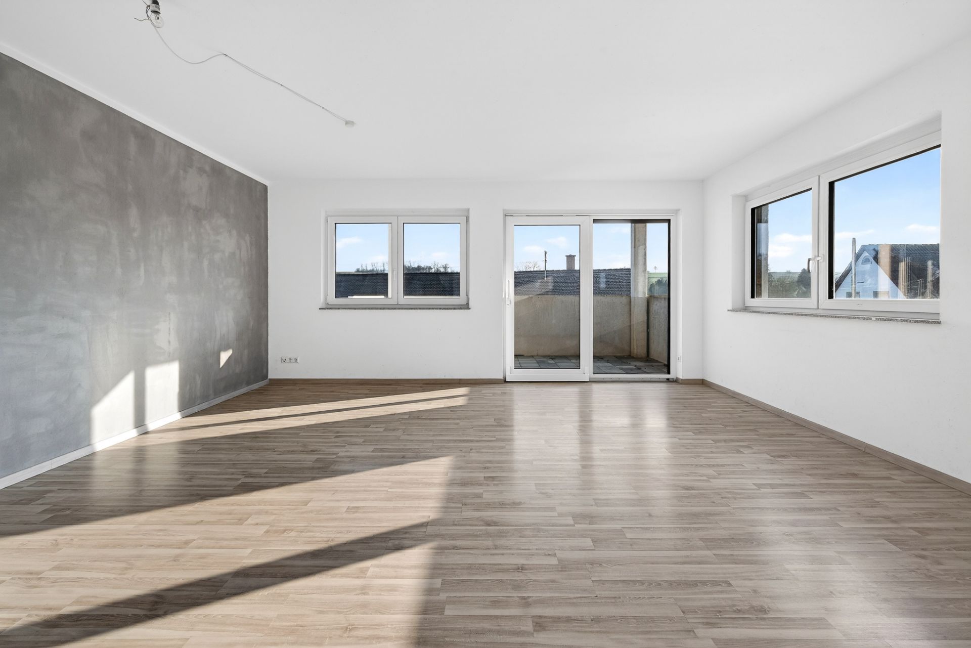 Helles, leeres Wohnzimmer mit Holzboden, großen Fenstern und Zugang zu einem Balkon.