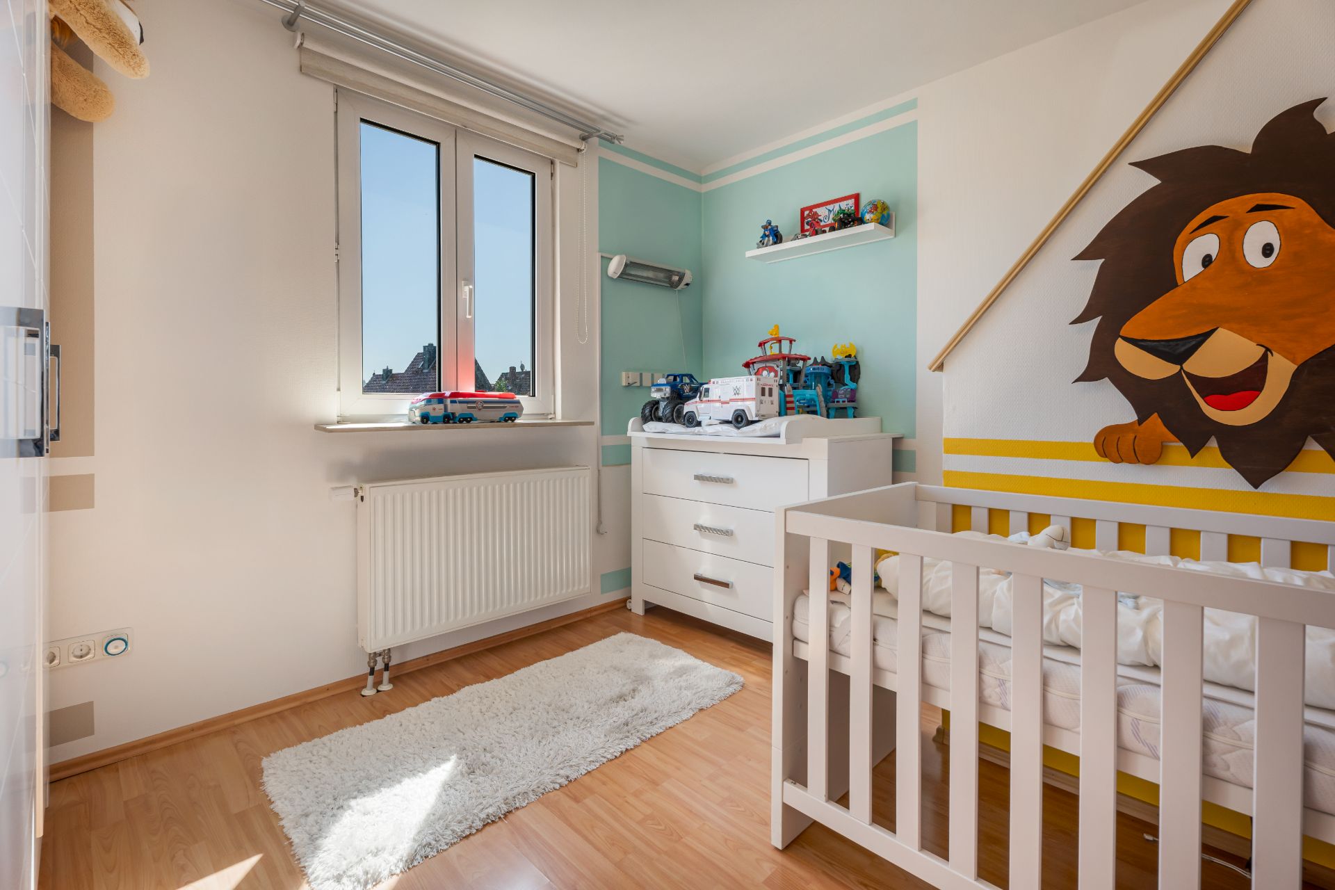 Helles Kinderzimmer mit Kinderbett, Teppich, Kommode und Löwenmotiv an der Wand.