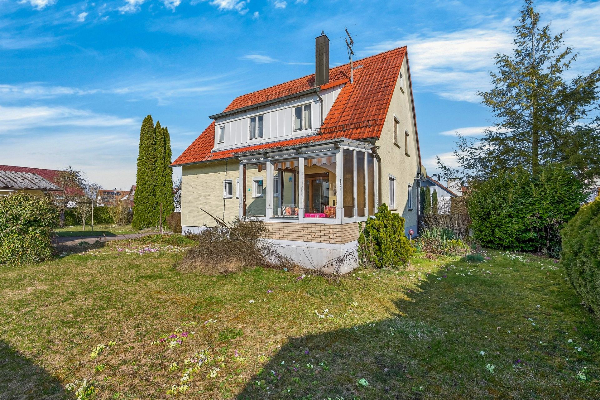 Zweistöckiges Haus mit rotem Dach und Wintergarten, umgeben von Garten und Bäumen.