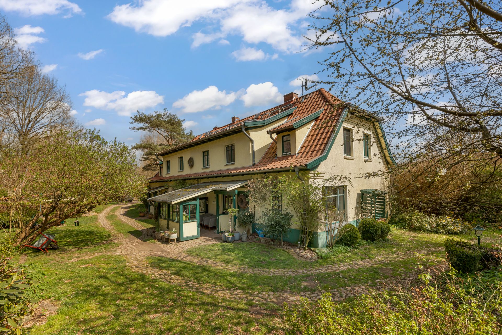 Zweistöckiges Haus mit rotem Dach und Garten, umgeben von Bäumen und Weg im Grünen.