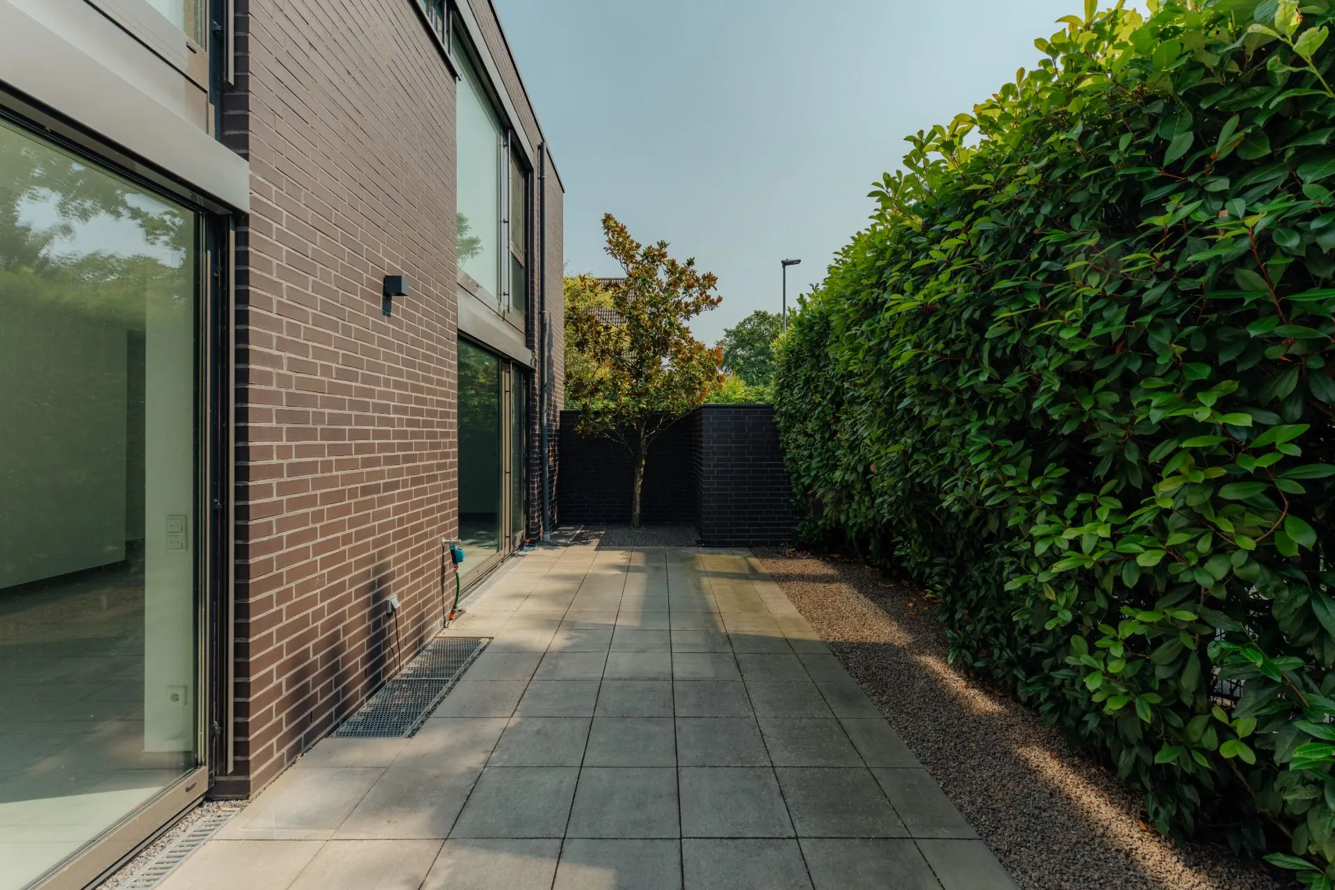 Gepflasterter Weg neben einem modernen Wohnhaus mit hoher grüner Hecke.
