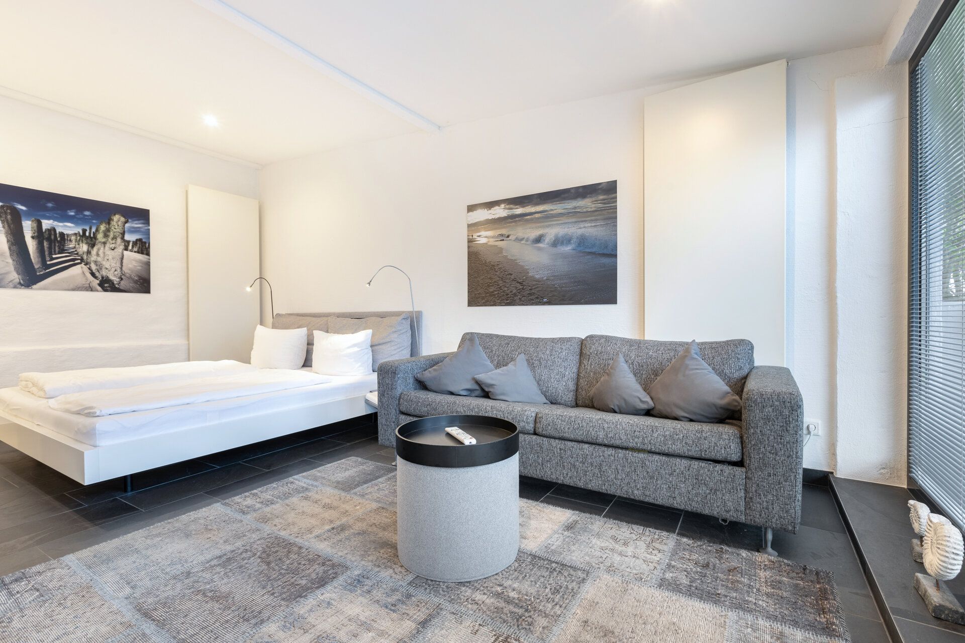 Helles Zimmer mit grauem Sofa, Bett, rundem Tisch und Bildern an weißen Wänden.