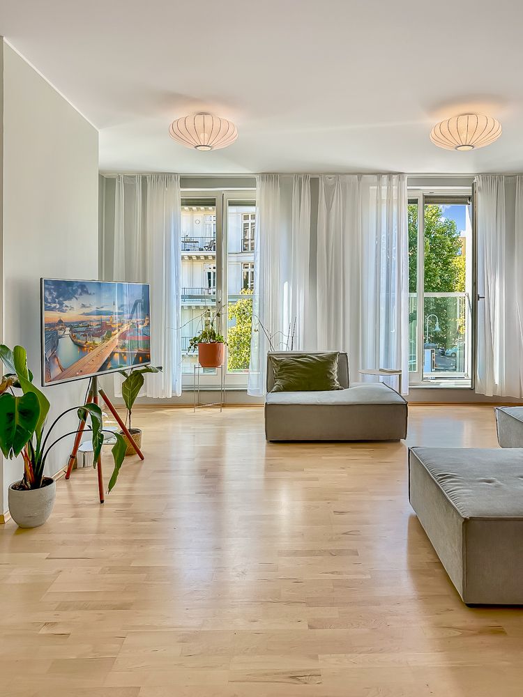 Helles Wohnzimmer mit großen Fenstern, Holzboden, Sofa, Fernseher und Zimmerpflanzen.
