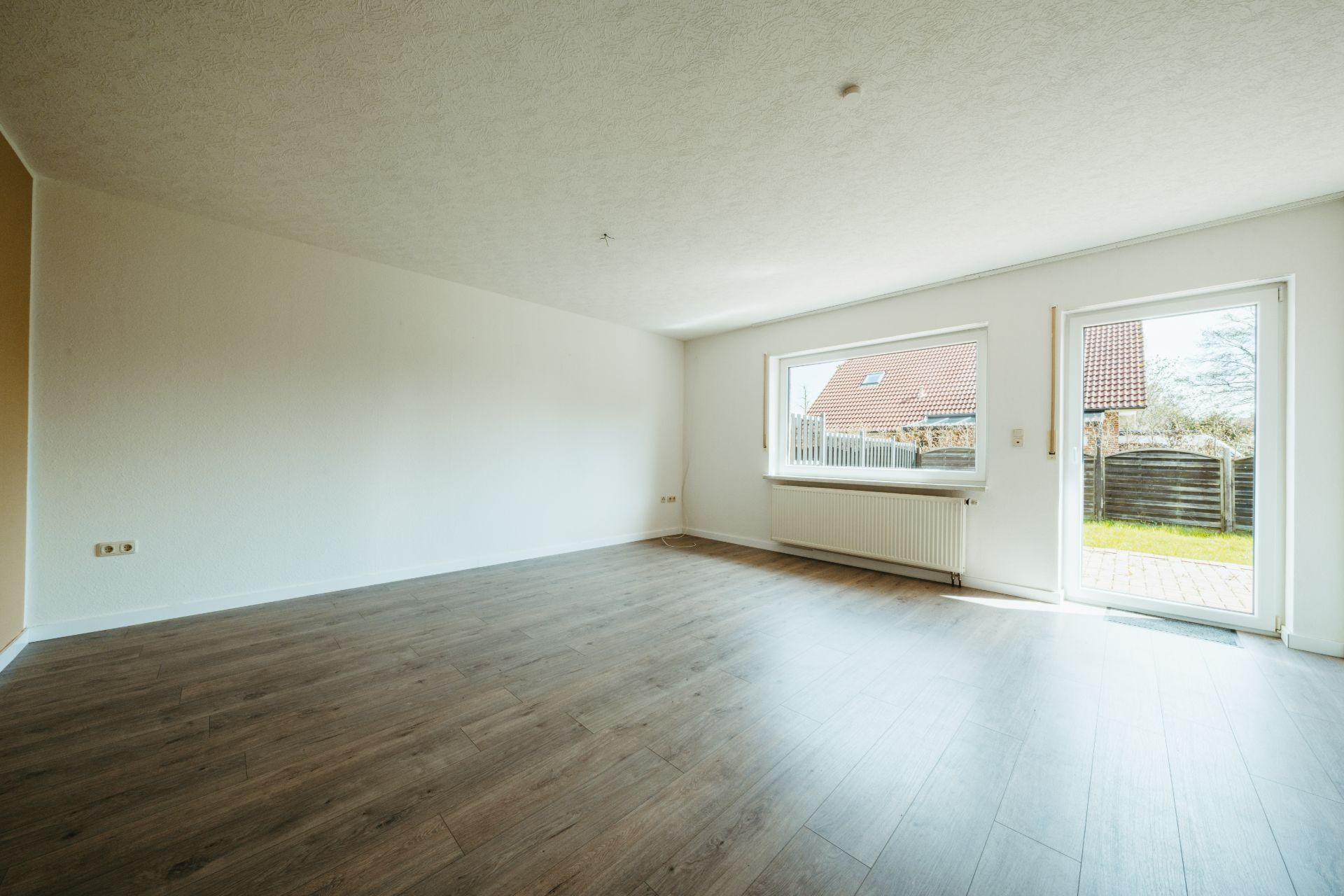 Helles, leeres Zimmer mit Holzfußboden, großem Fenster und Tür zum Garten.