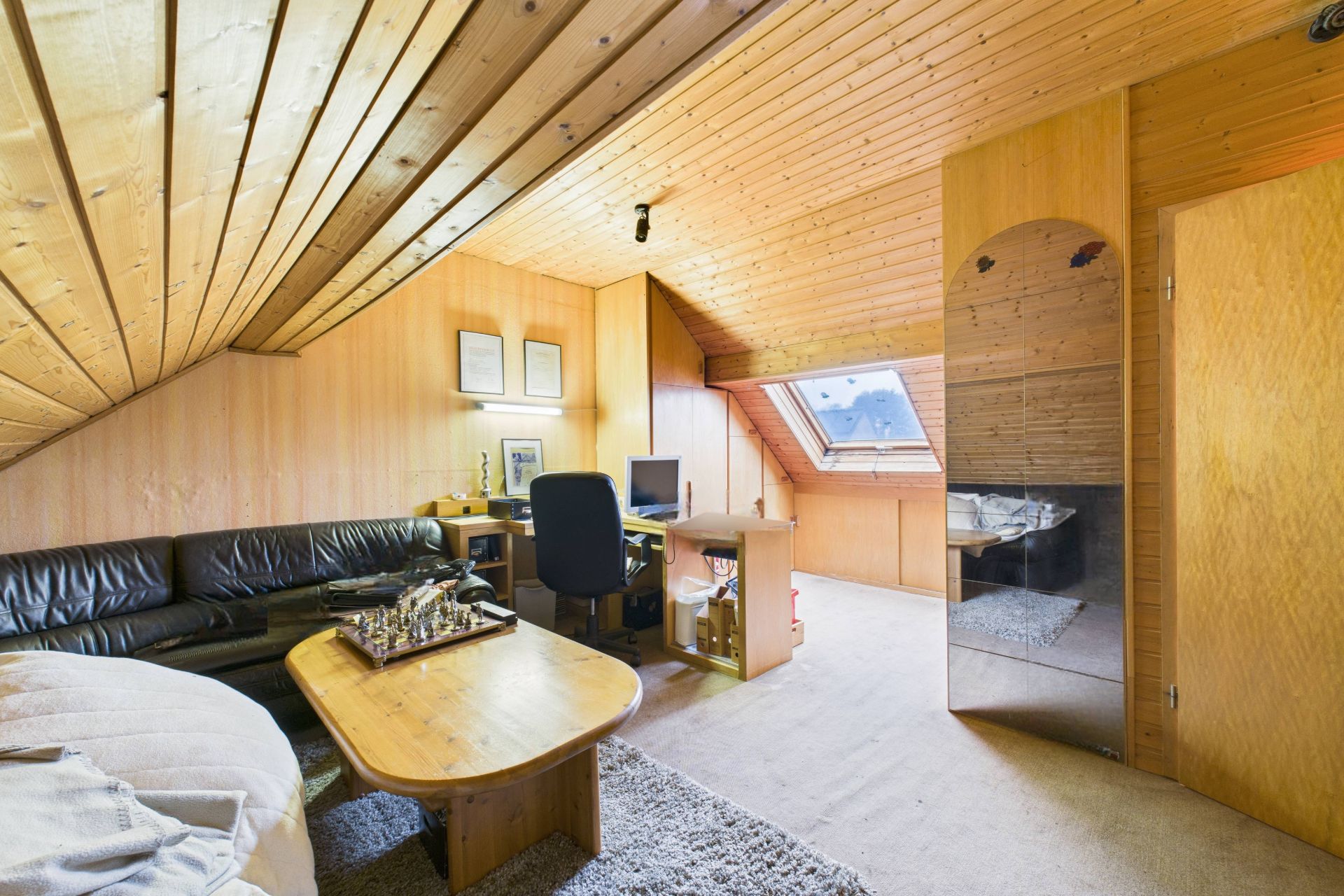 Wohnzimmer mit Holzdecke, Sofa, Schreibtisch, Dachfenster und Spiegel an der Wand.