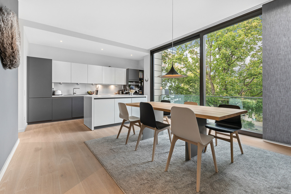 Moderne Küche mit Essbereich, großem Holztisch und vier Stühlen vor großen Fenstern mit Blick ins Grüne.