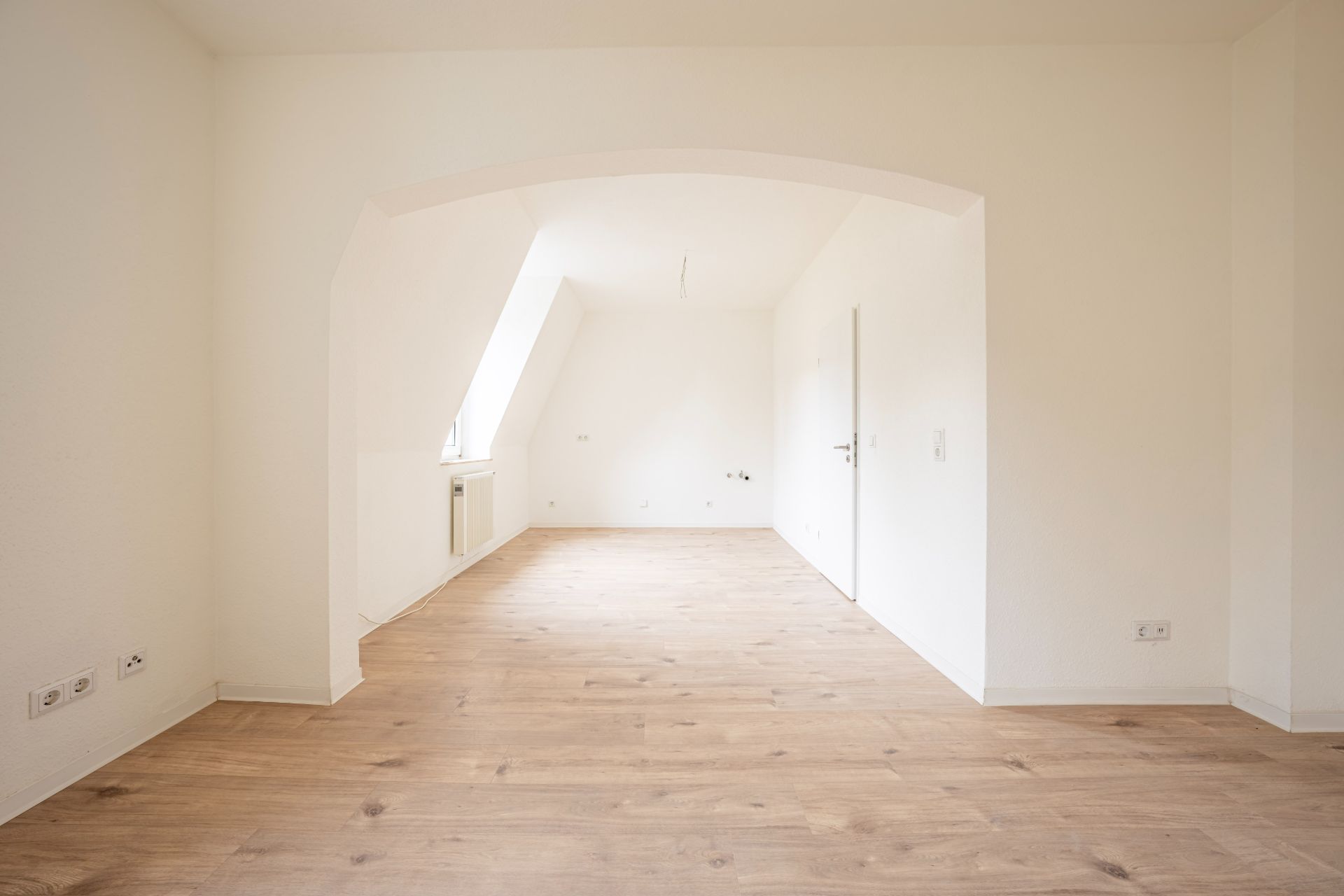 Heller leerer Raum mit Holzboden, weiß gestrichenen Wänden und einem Bogen in der Mitte.