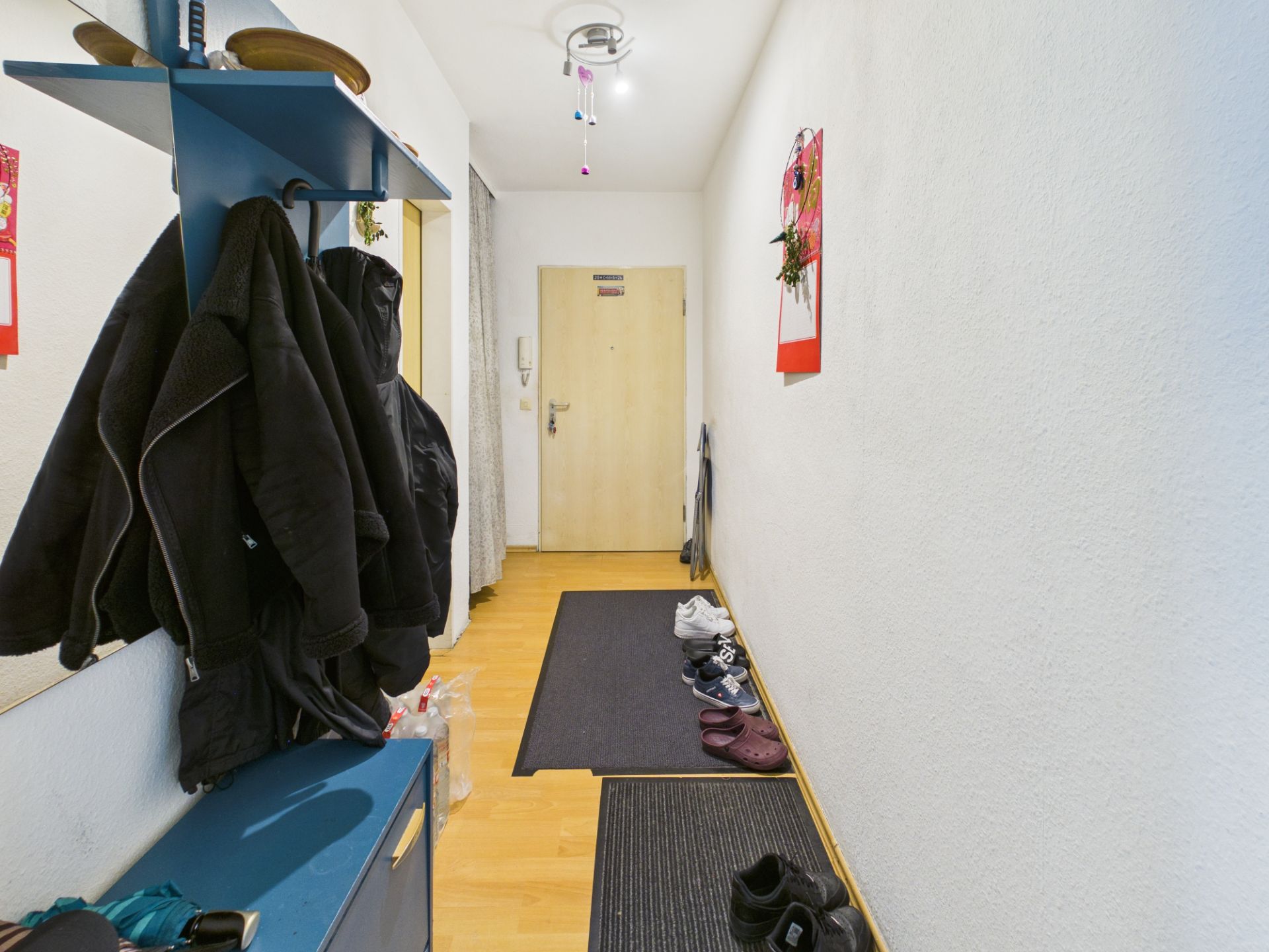 Flur mit Garderobe, mehreren Jacken, Schuhen auf Matten und heller Holztür am Ende.
